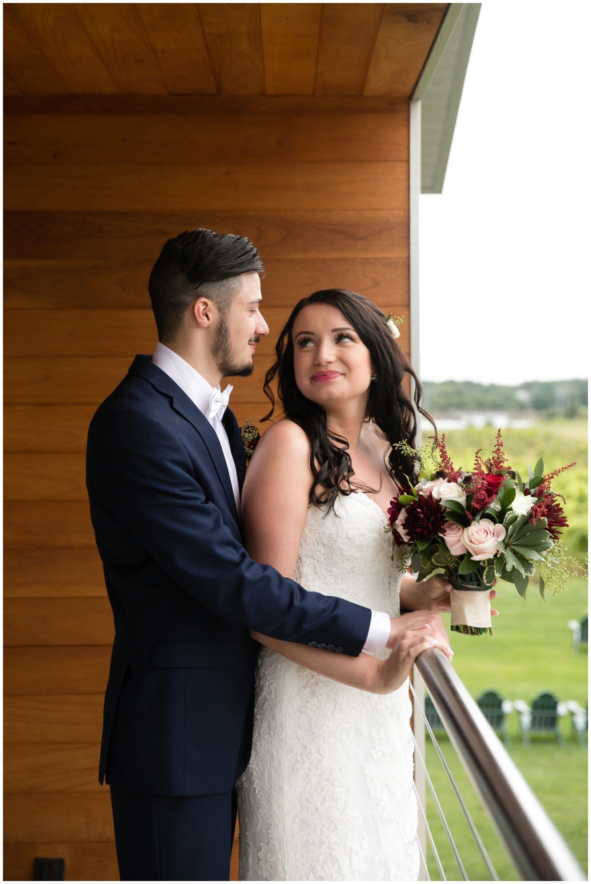 Bride and groom in navy and white wedding attire standing on a balcony, holding a bouquet and gazing at each other.