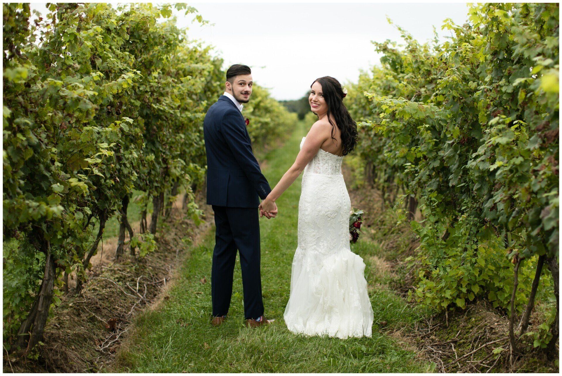 Couple holding hands in a vineyard aisle, wearing wedding attire and looking back at the camera.