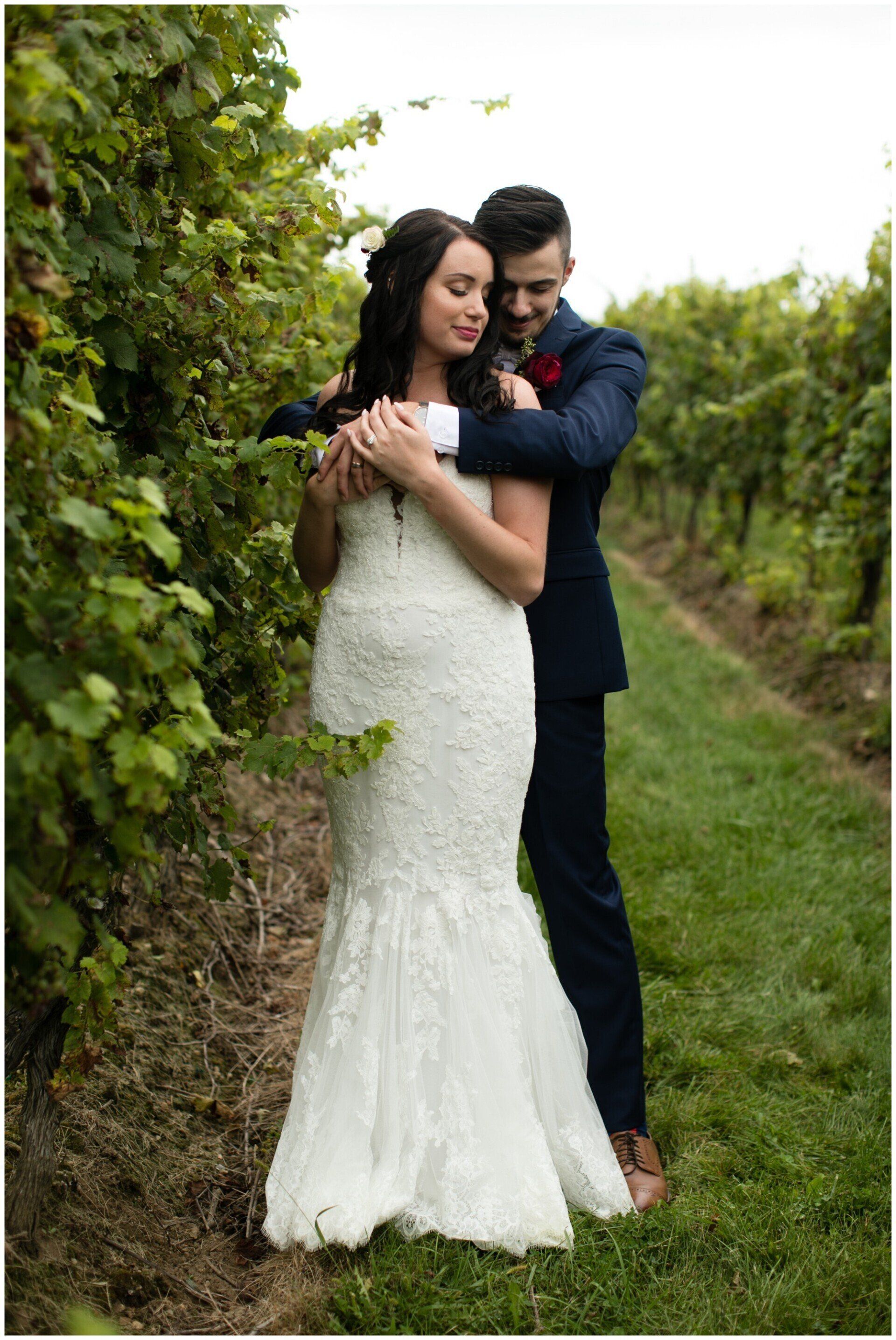 Bride and groom embracing in a vineyard, standing on a grassy path beside leafy vines.