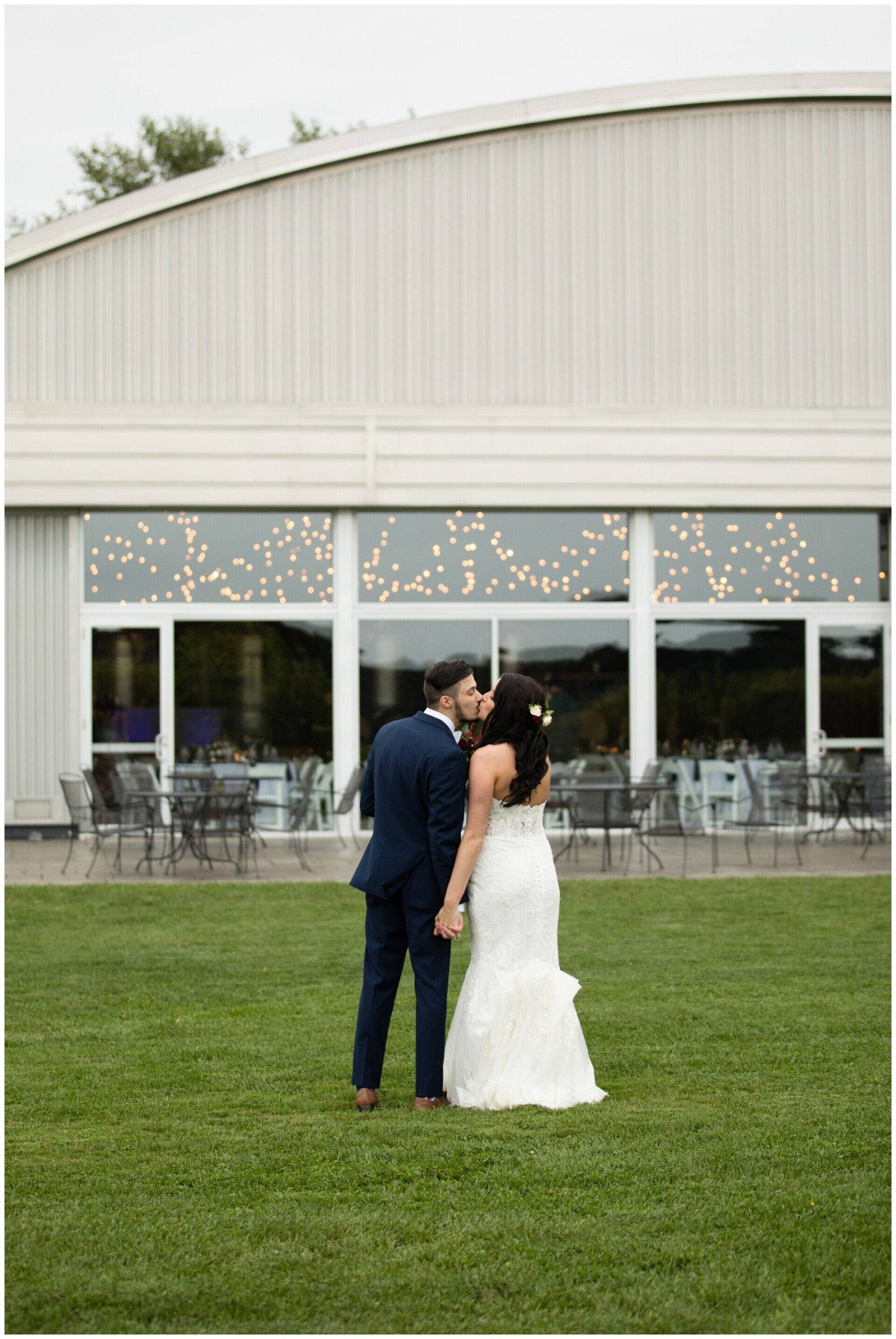 Newlyweds standing on a grassy lawn in front of a white event hall, sharing a kiss