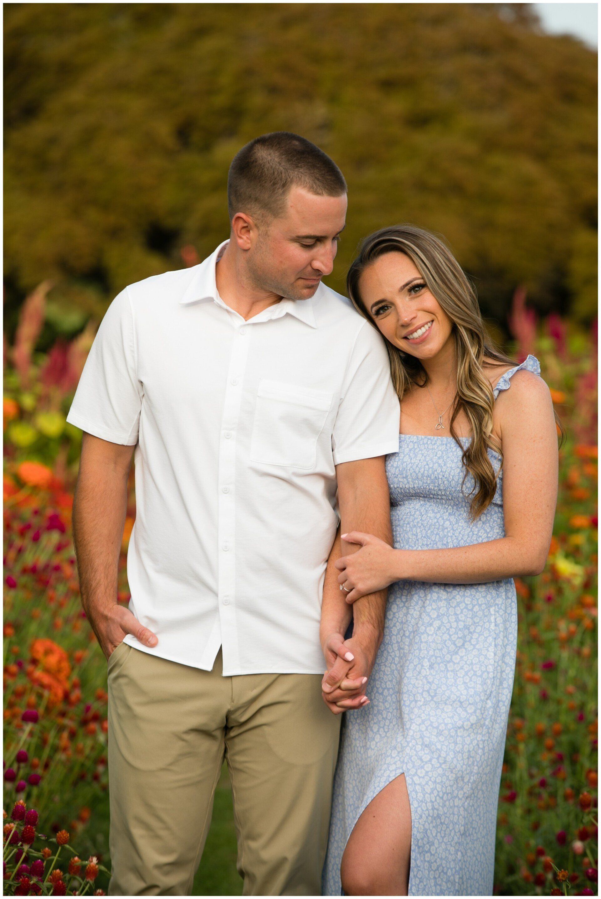 Couple standing in a flower field, holding hands and leaning together in a soft outdoor portrait.