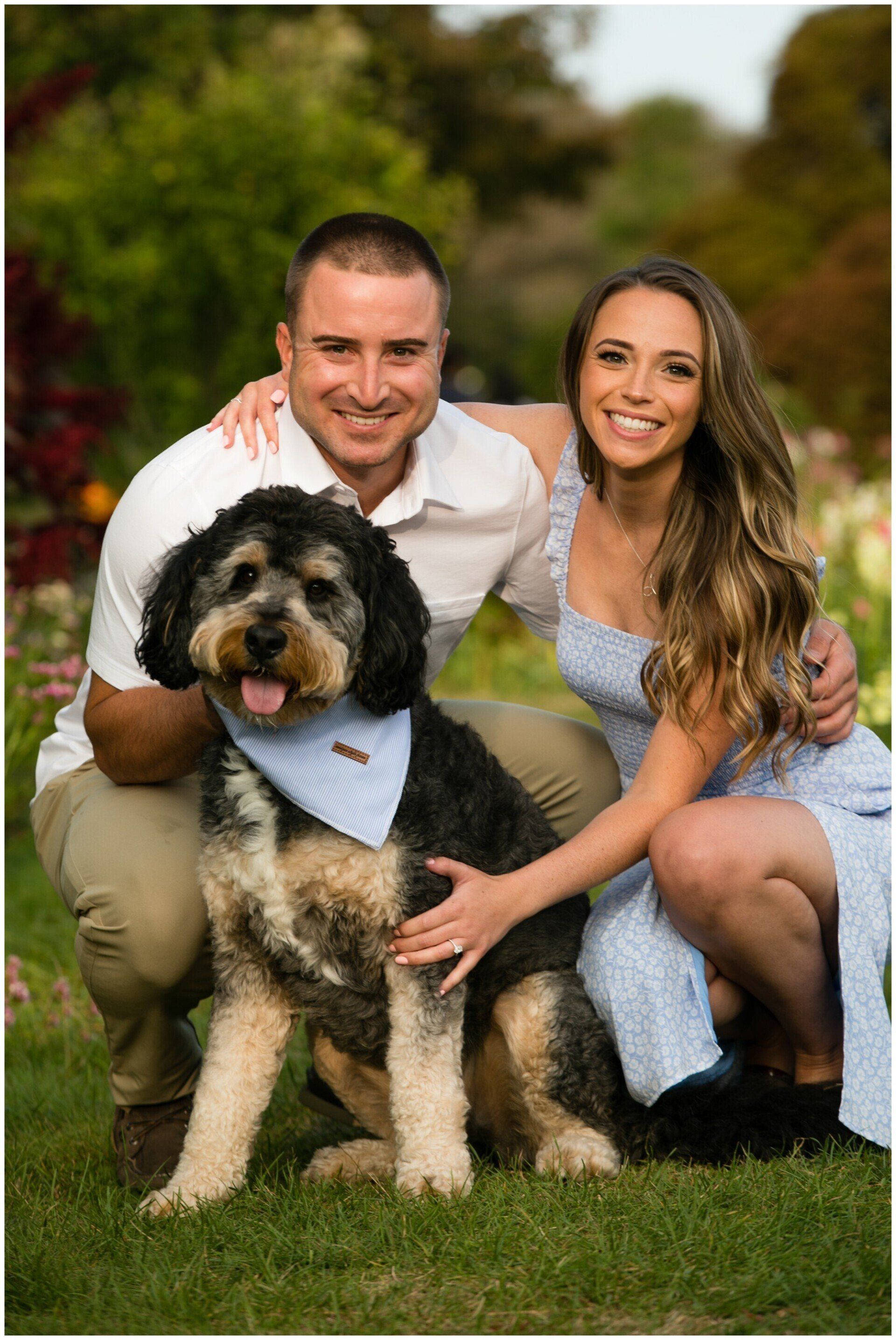 Smiling couple crouches beside a large black-and-tan dog on a grassy lawn outdoors.