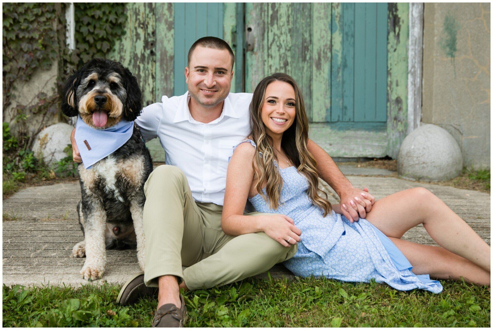 Couple sitting on grass with a dog in front of a rustic door, smiling outdoors