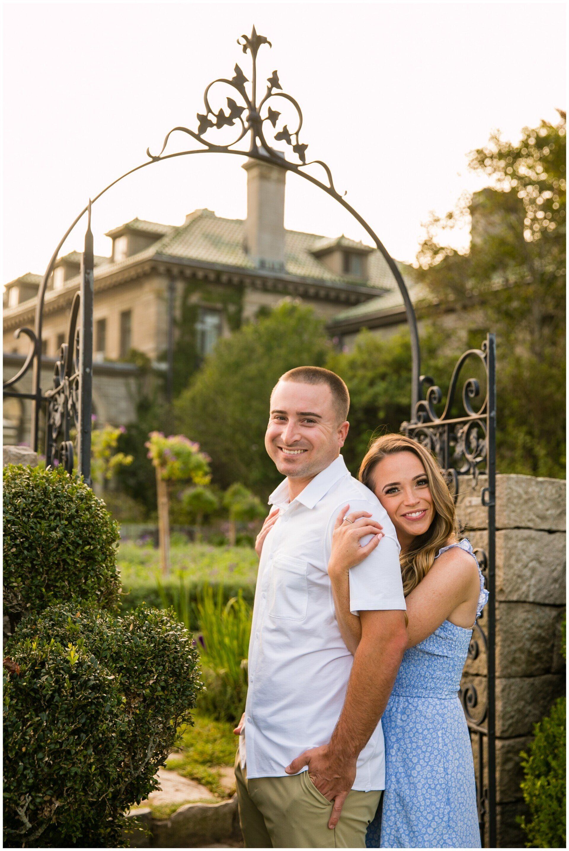 Couple posing outdoors in front of a mansion and garden gate, smiling at the camera.