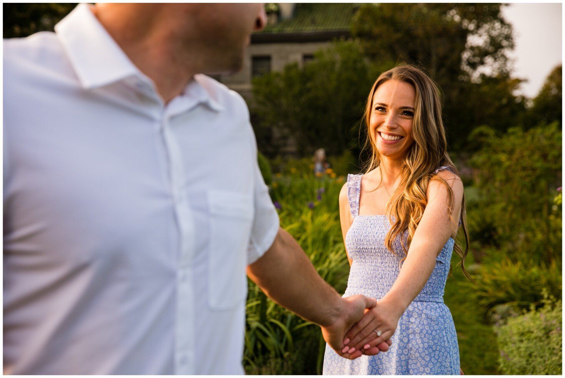 Smiling woman in a white dress holding hands with a man in a white shirt outdoors in a garden