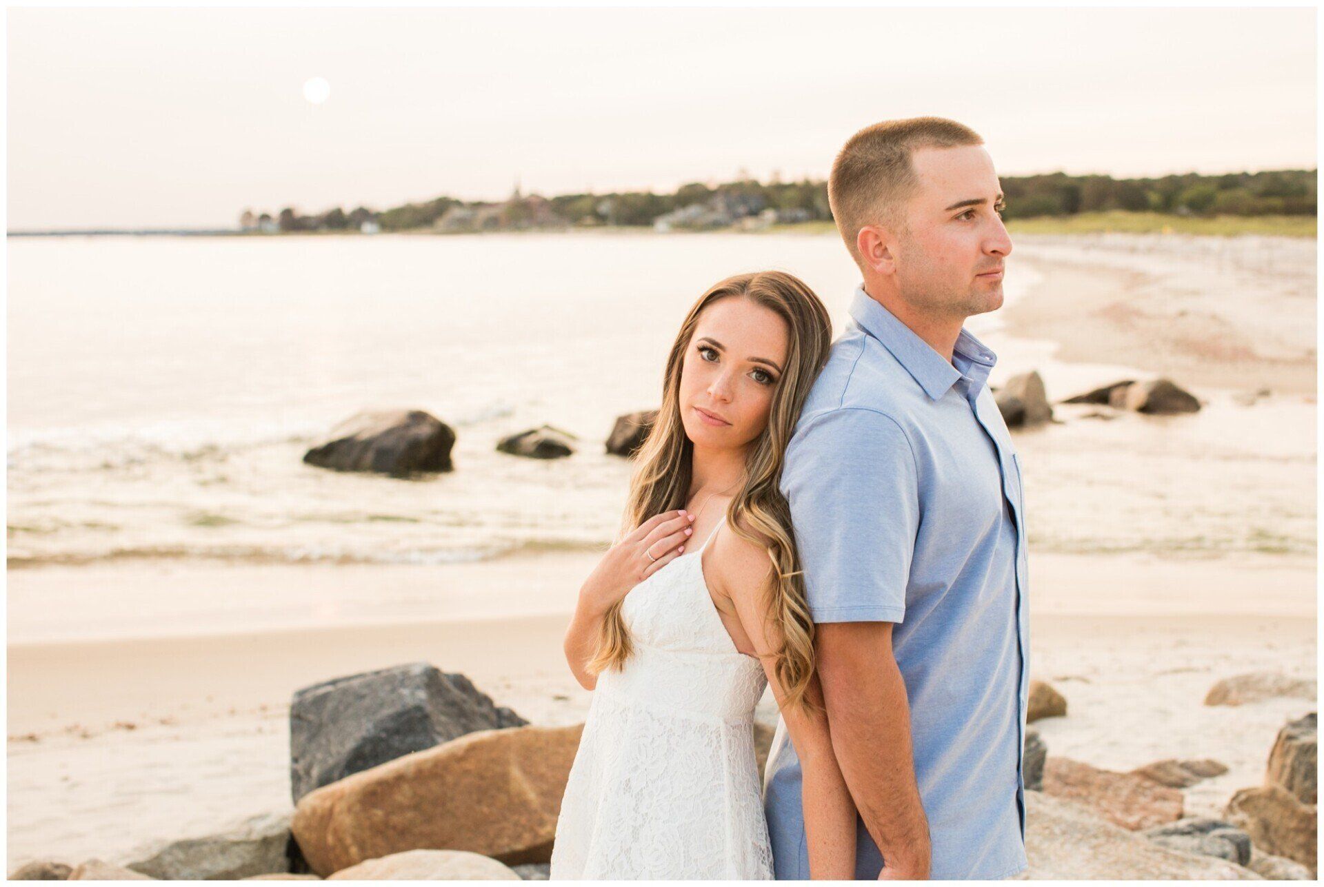 Couple standing back-to-back on a rocky beach at sunset, looking in opposite directions.