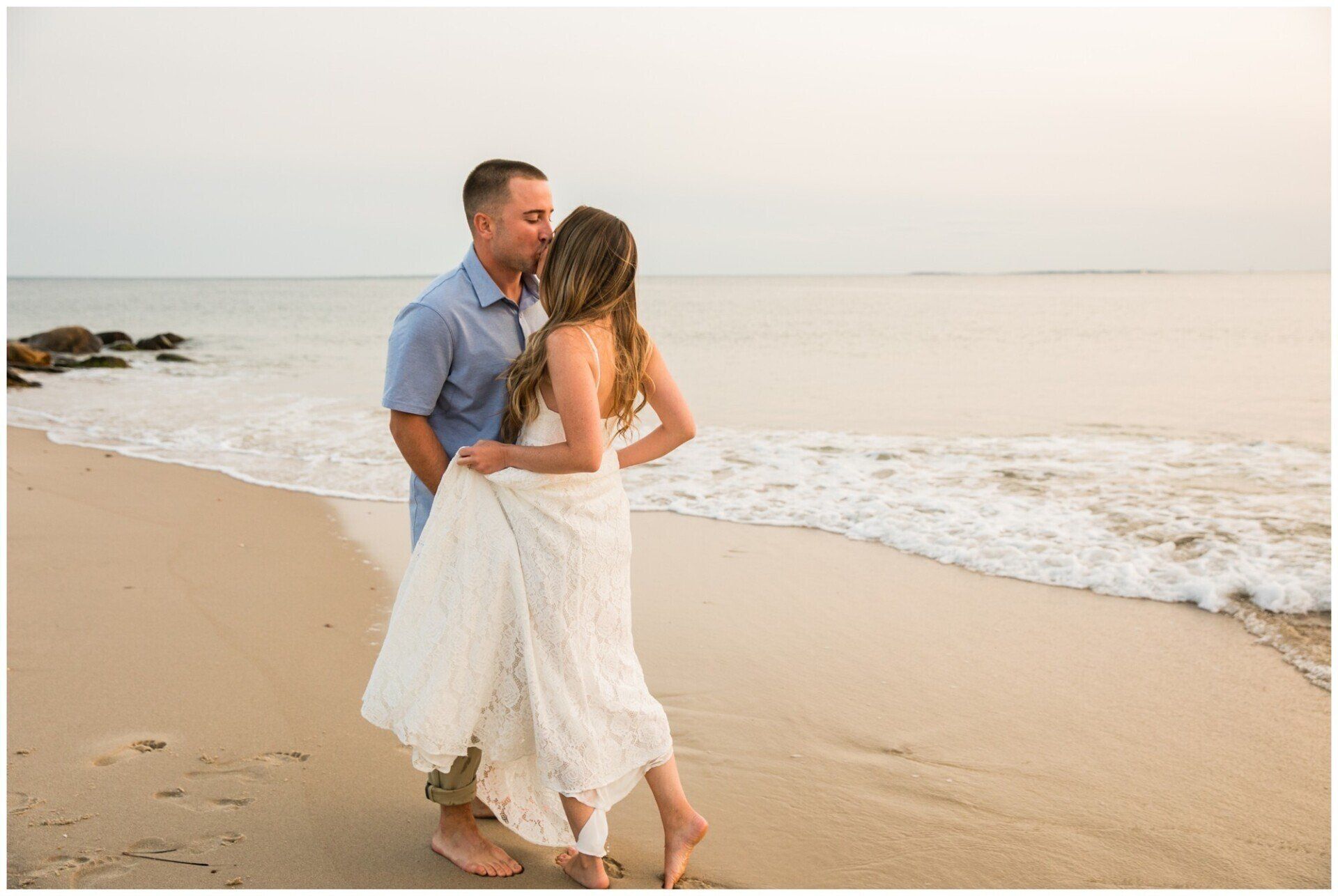 Couple walking barefoot on a sandy beach at sunset, facing the calm ocean.