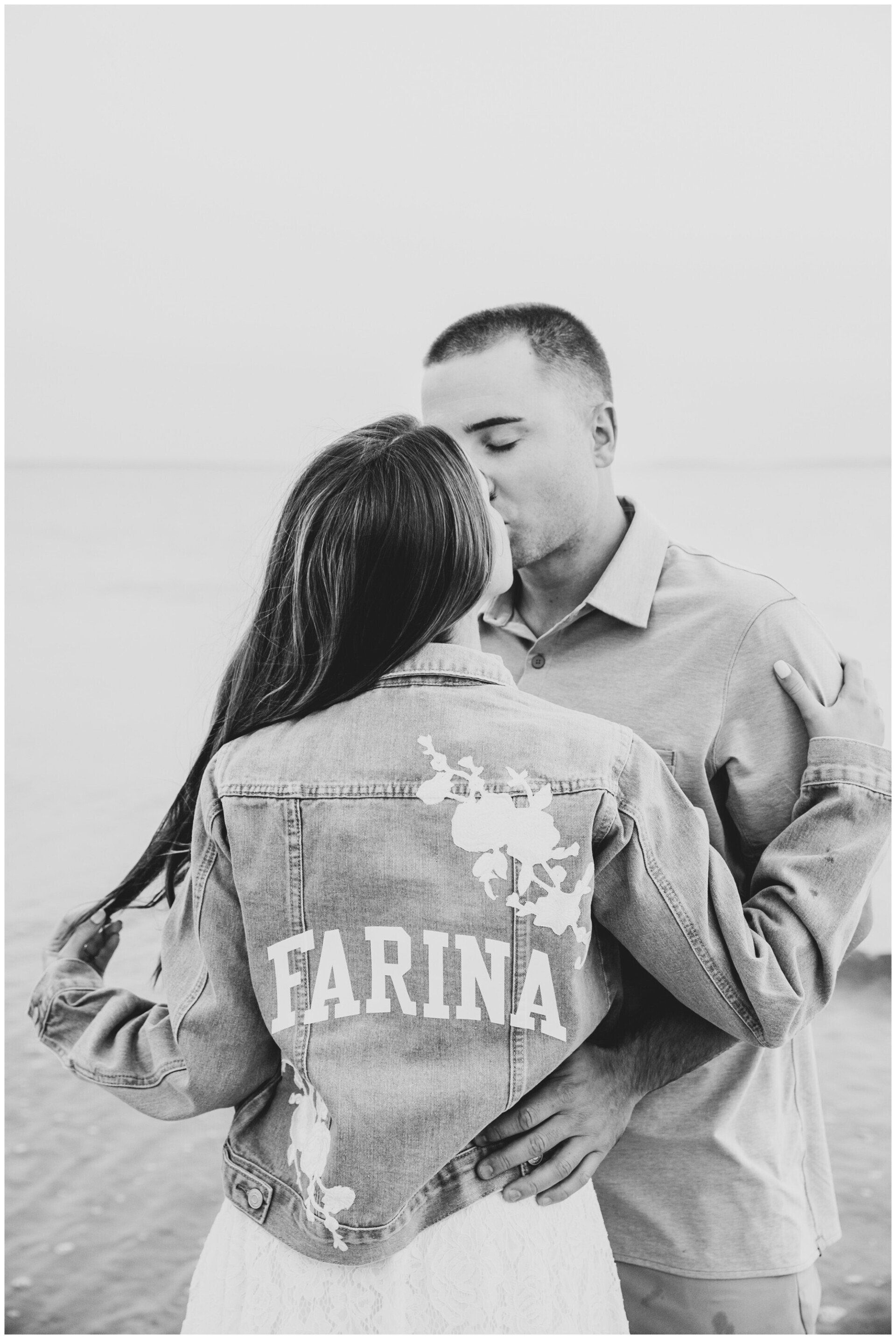 Couple embracing on a beach, woman in denim jacket with “FARINA” on the back.