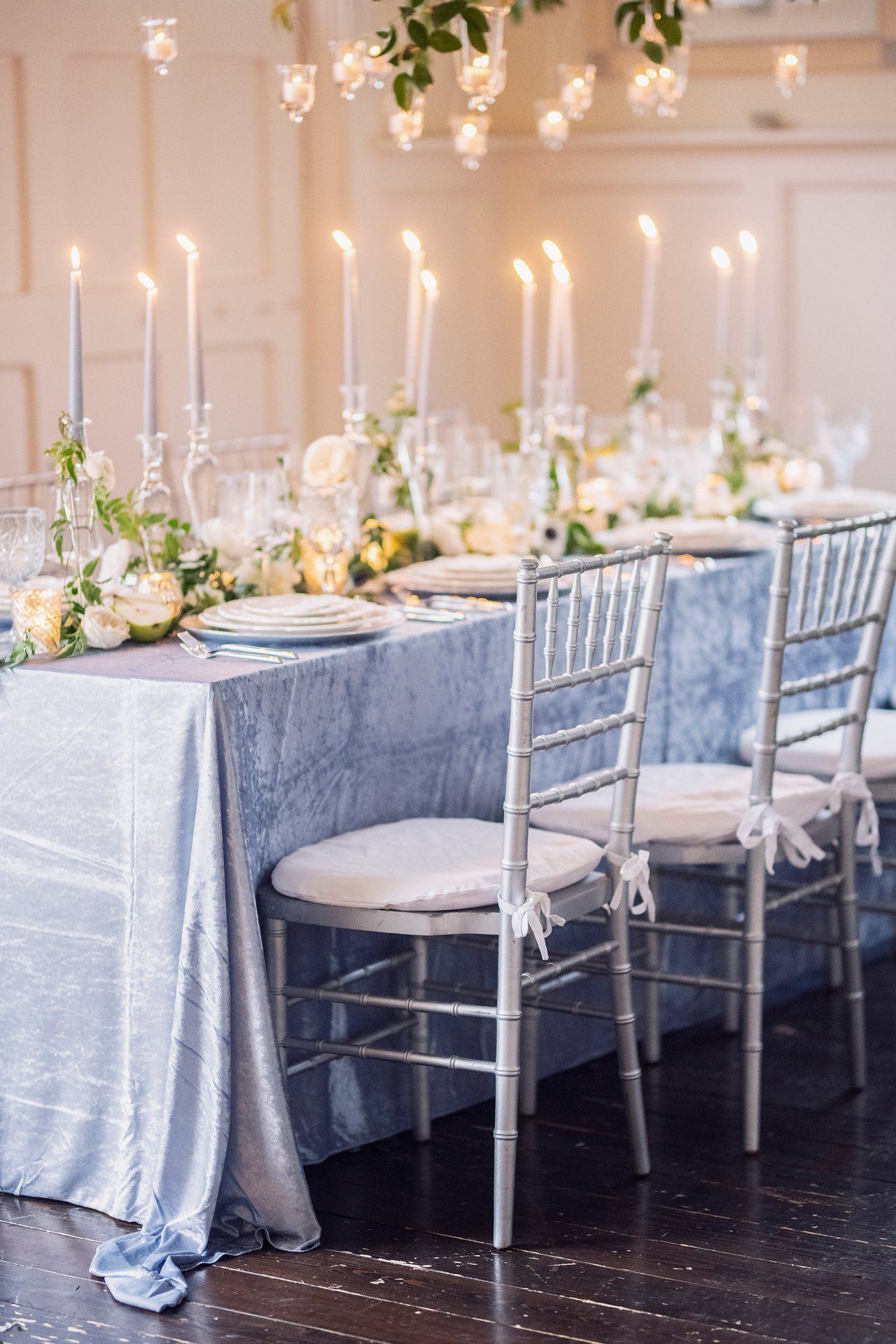 Elegant candlelit dining table with white flowers, silver chairs, and pale blue linens in a formal reception room