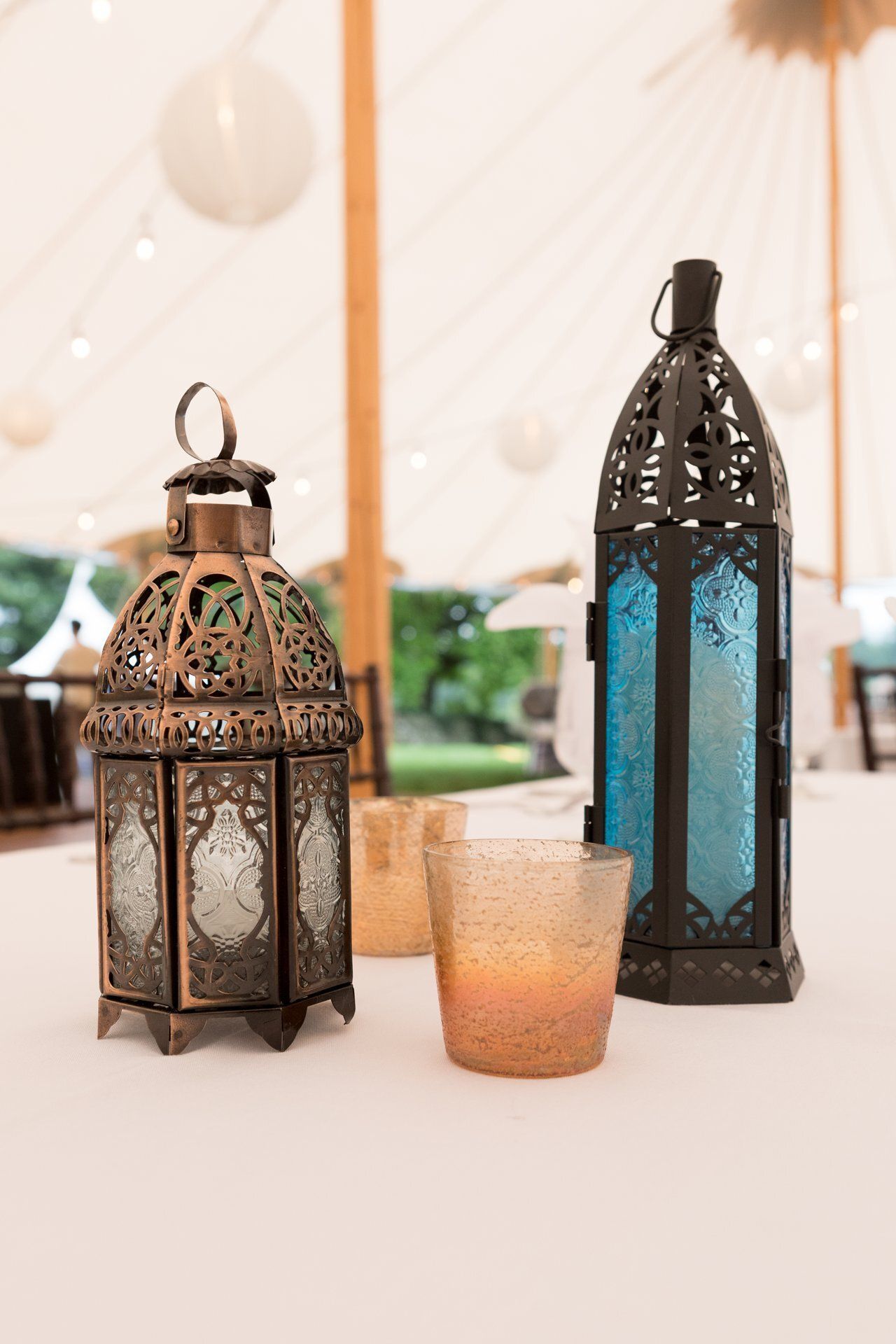 Decorative lanterns and a glass candle holder on a white table under a bright tent.