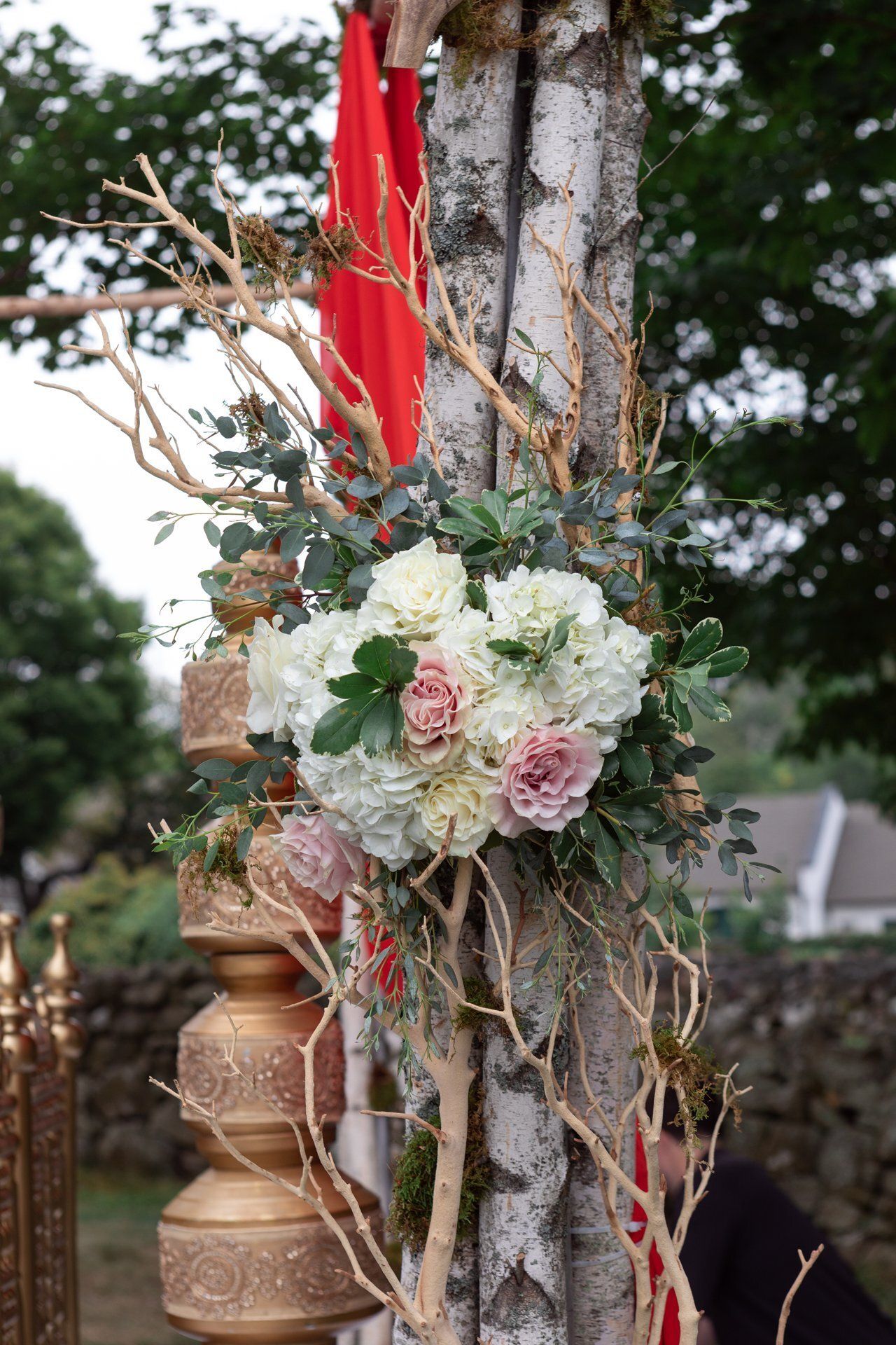 Floral wreath with white and blush flowers tied to a birch tree beside red ribbons and decorative vessels