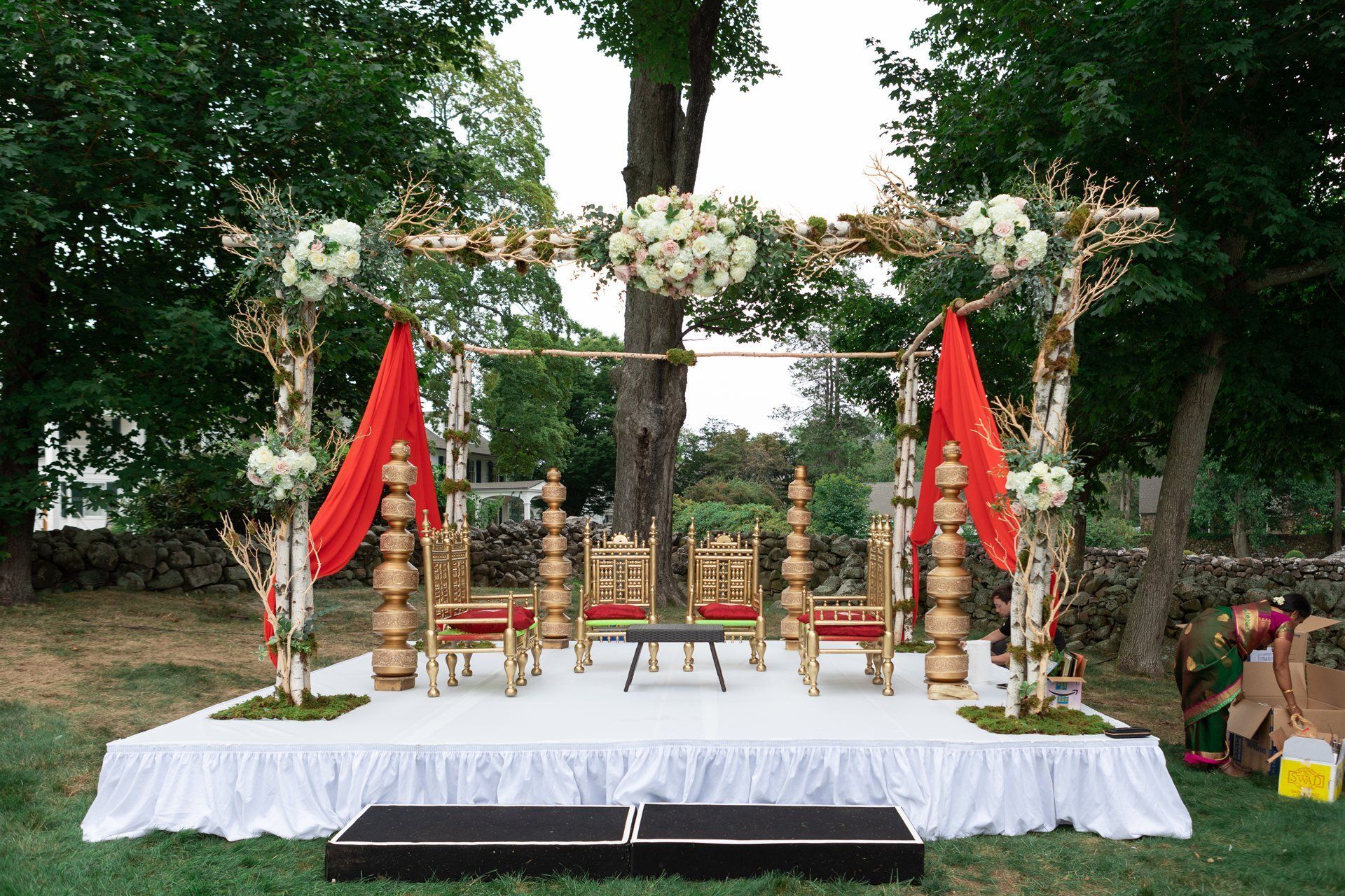 Decorated outdoor ceremonial stage with white canopy, red flags, brass ornaments, and chairs on a lawn