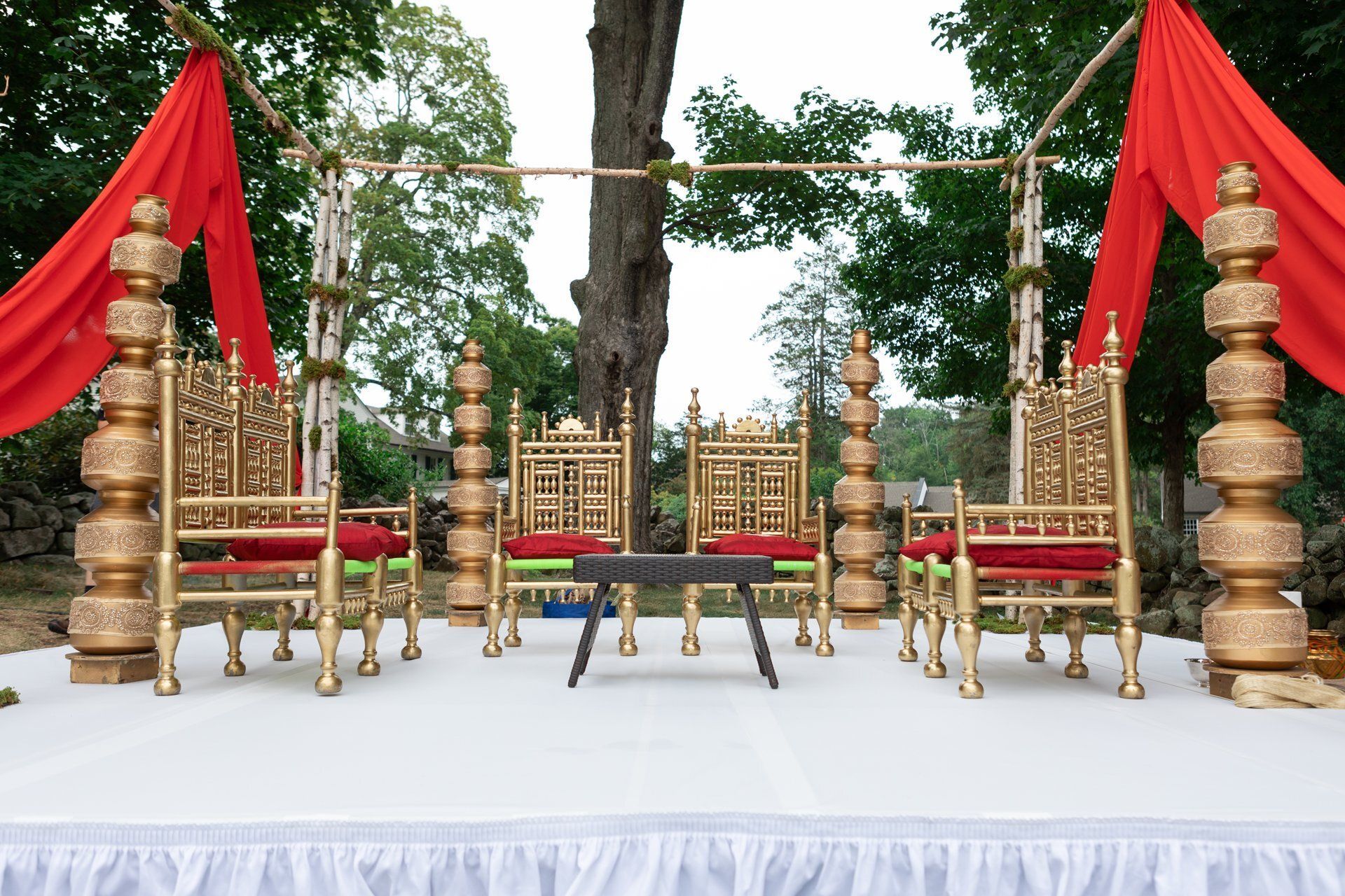 Decorated outdoor stage with gold chairs, red drapes, and white tablecloths under trees
