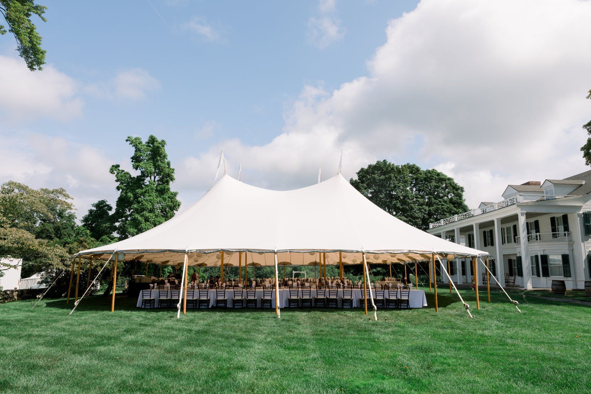 Large white event tent on a green lawn with people seated underneath, beside a white colonial-style building.