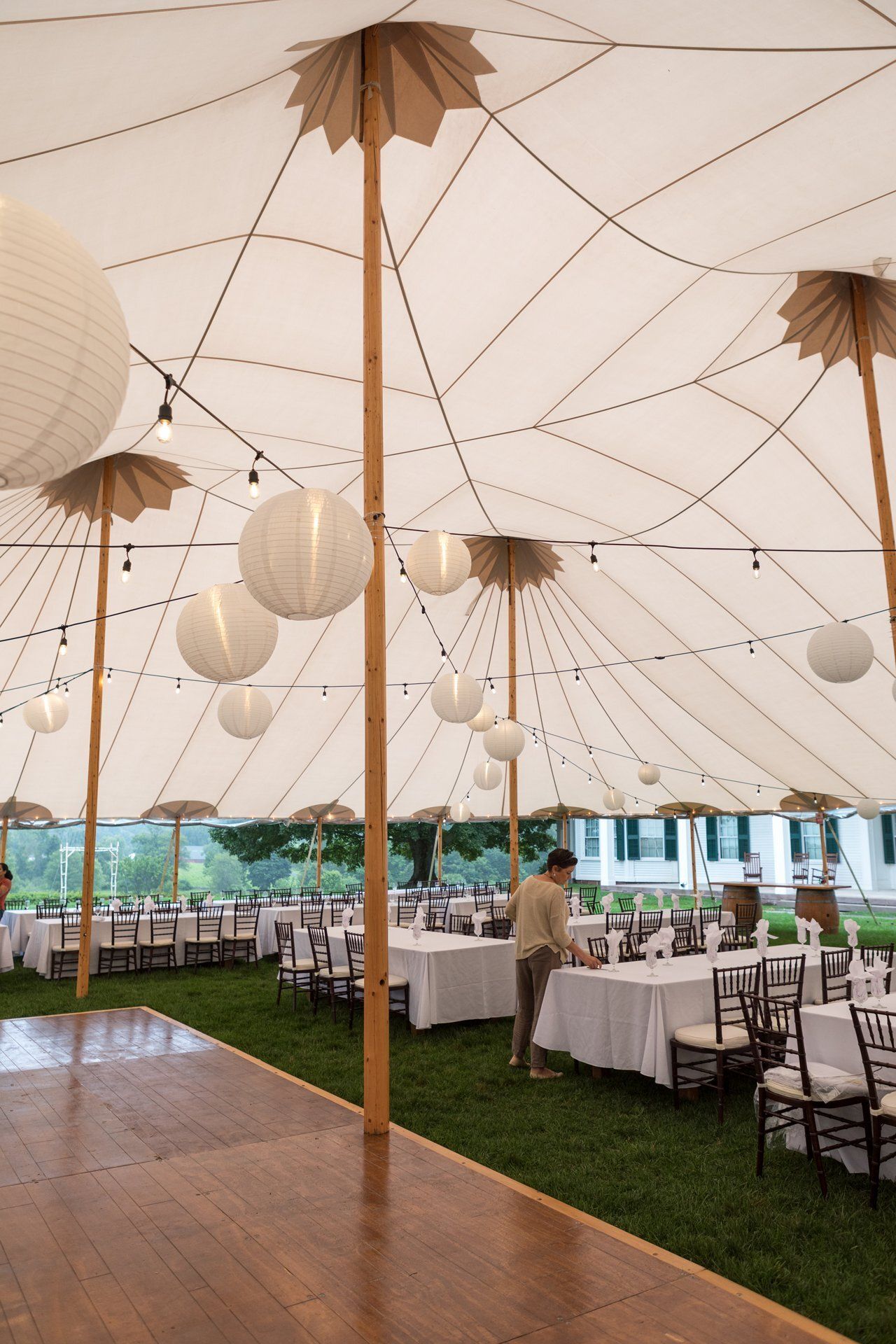 White-tented outdoor dining setup with hanging lanterns, long tables, and green carpet under a peaked ceiling