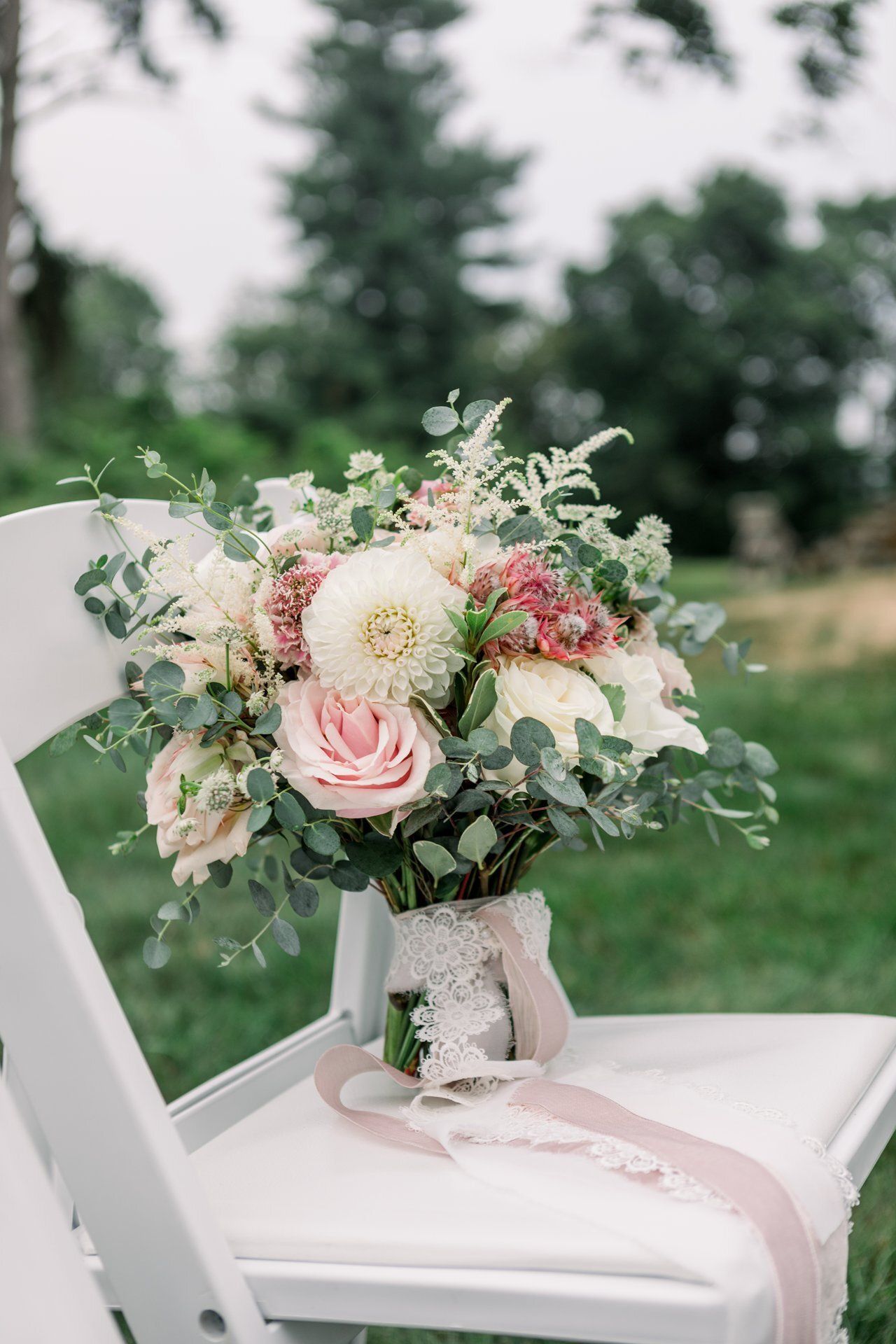 Pastel wedding bouquet on a white chair outdoors, with soft pink ribbon and greenery