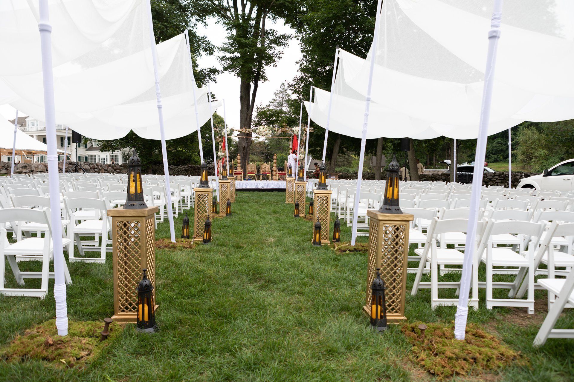 Outdoor wedding aisle with white chairs and canopies on a grassy lawn, lined with lanterns.