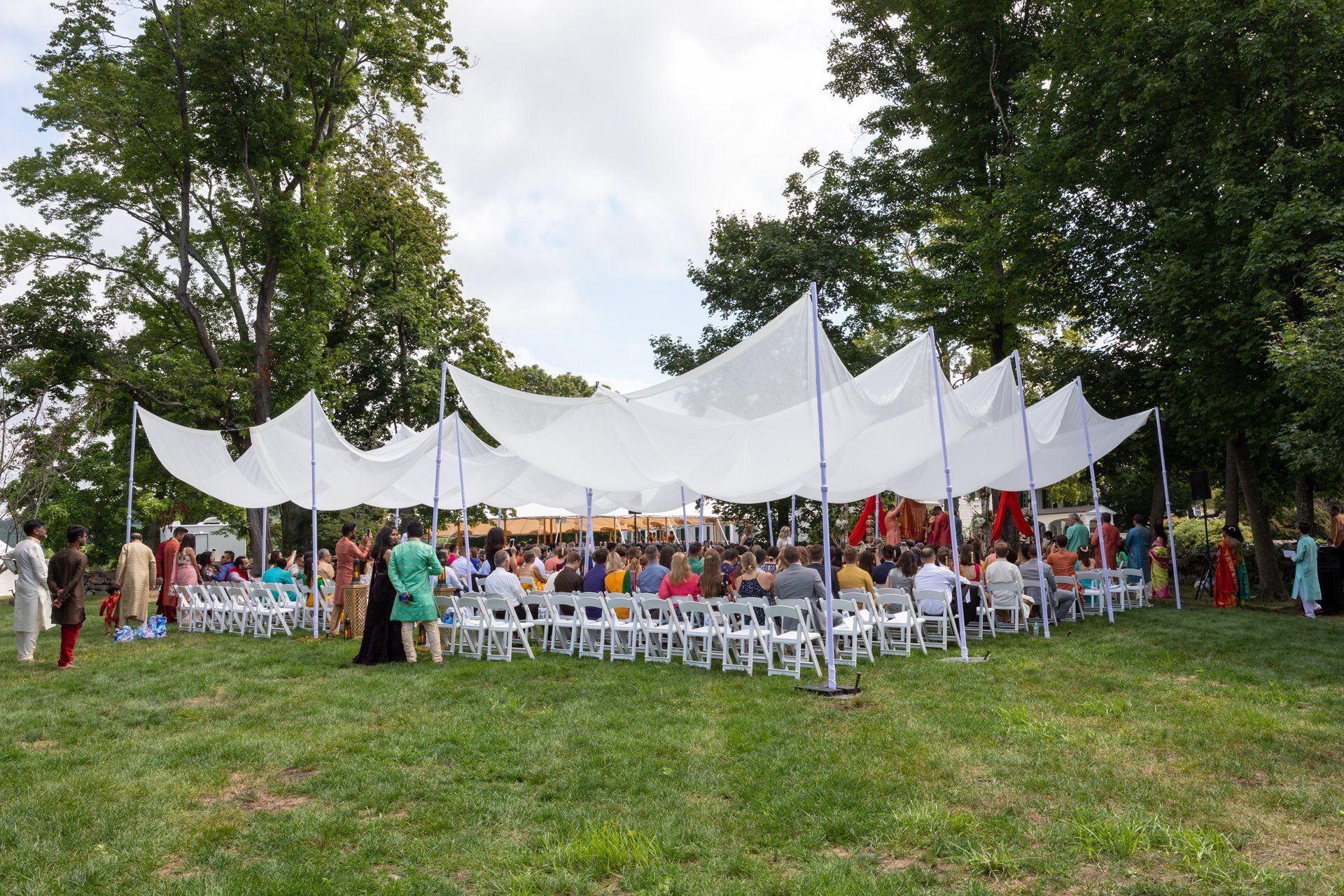 Outdoor gathering under white tents with seated guests on a grassy lawn surrounded by trees