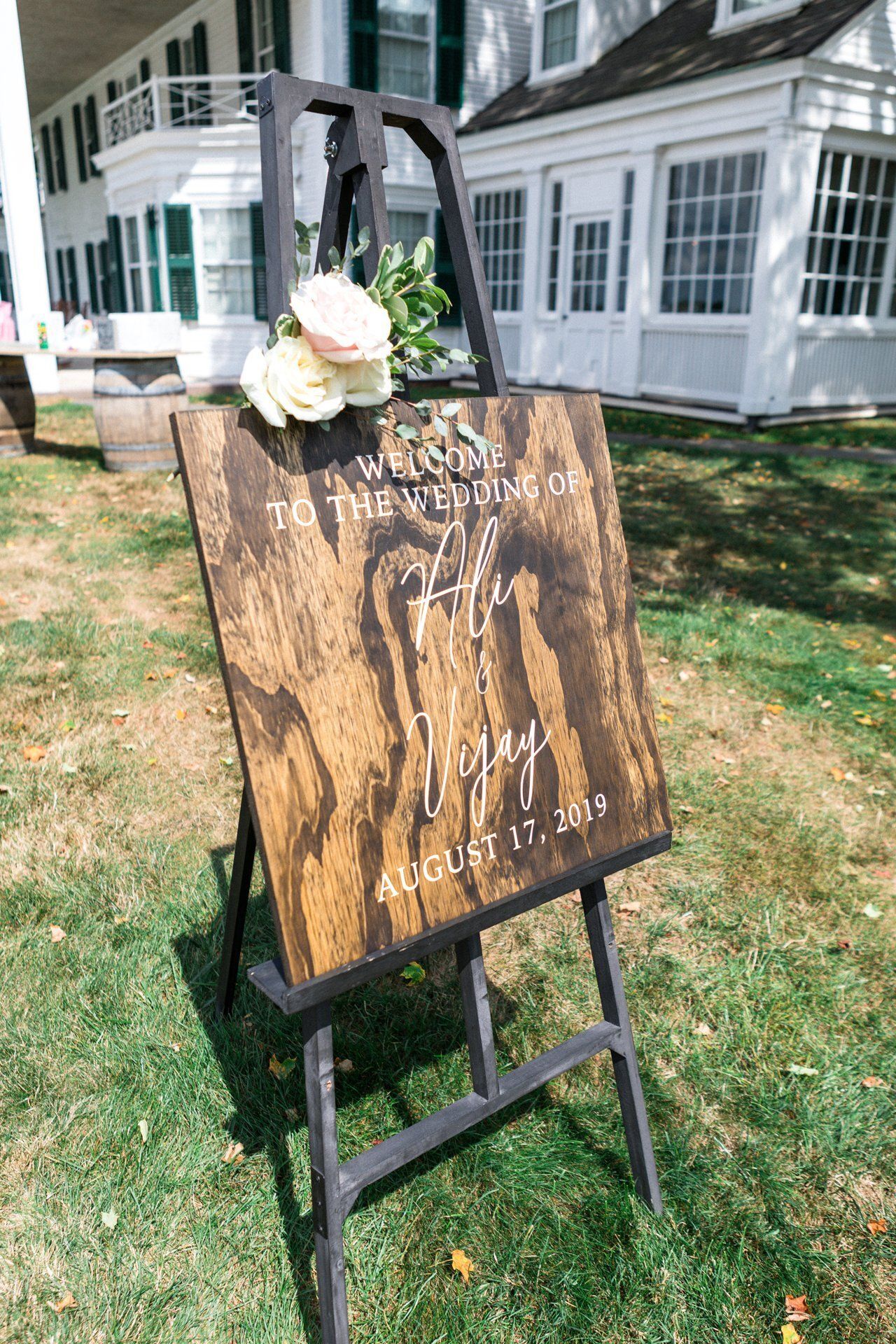 Rustic wooden sign on a black easel with white flowers, outside a white house on a grassy lawn.