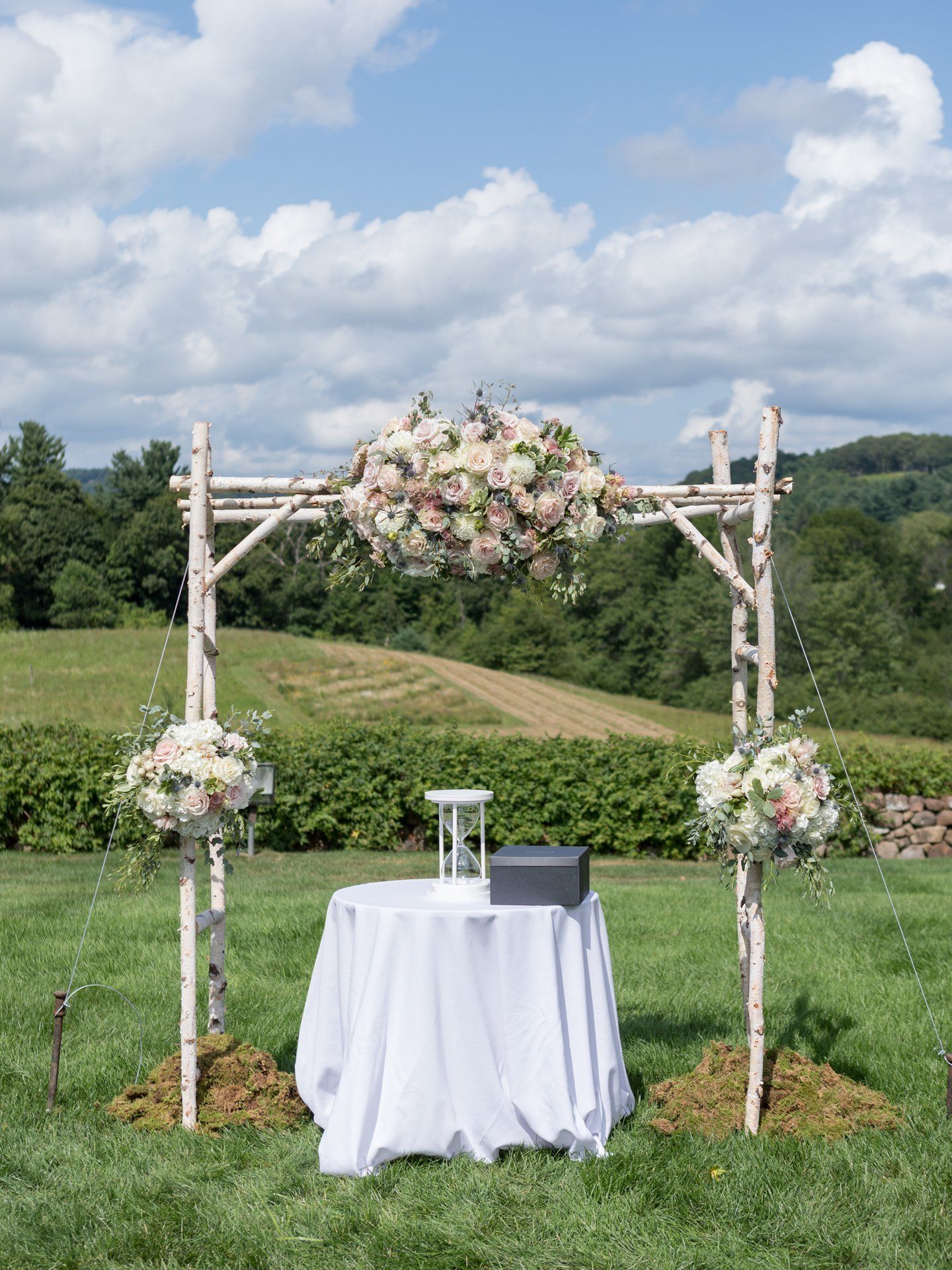 Outdoor wedding altar with white table and floral arch on a grassy field under a cloudy sky