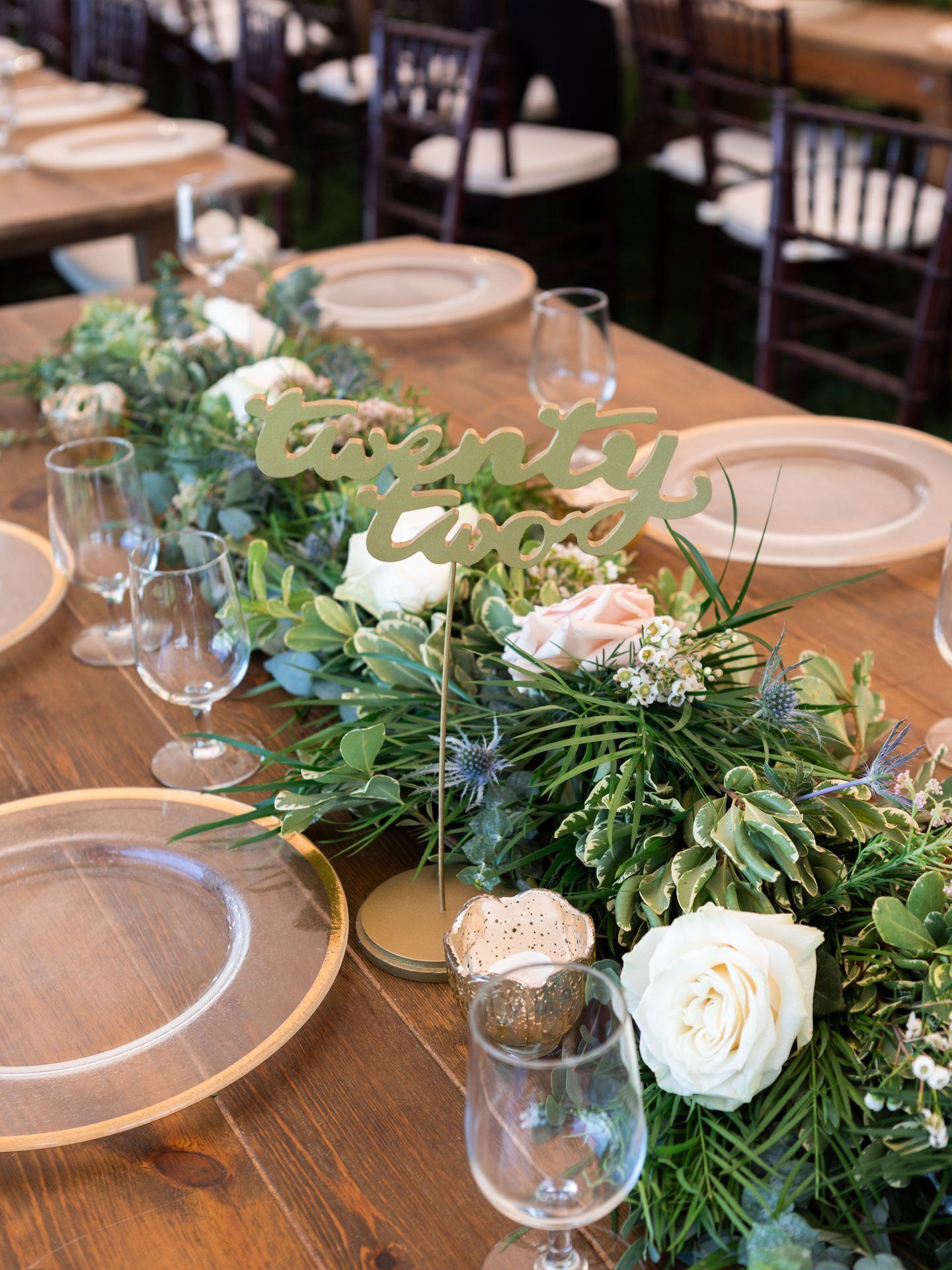 Wooden banquet table with greenery, white roses, glassware, and a gold “love” centerpiece
