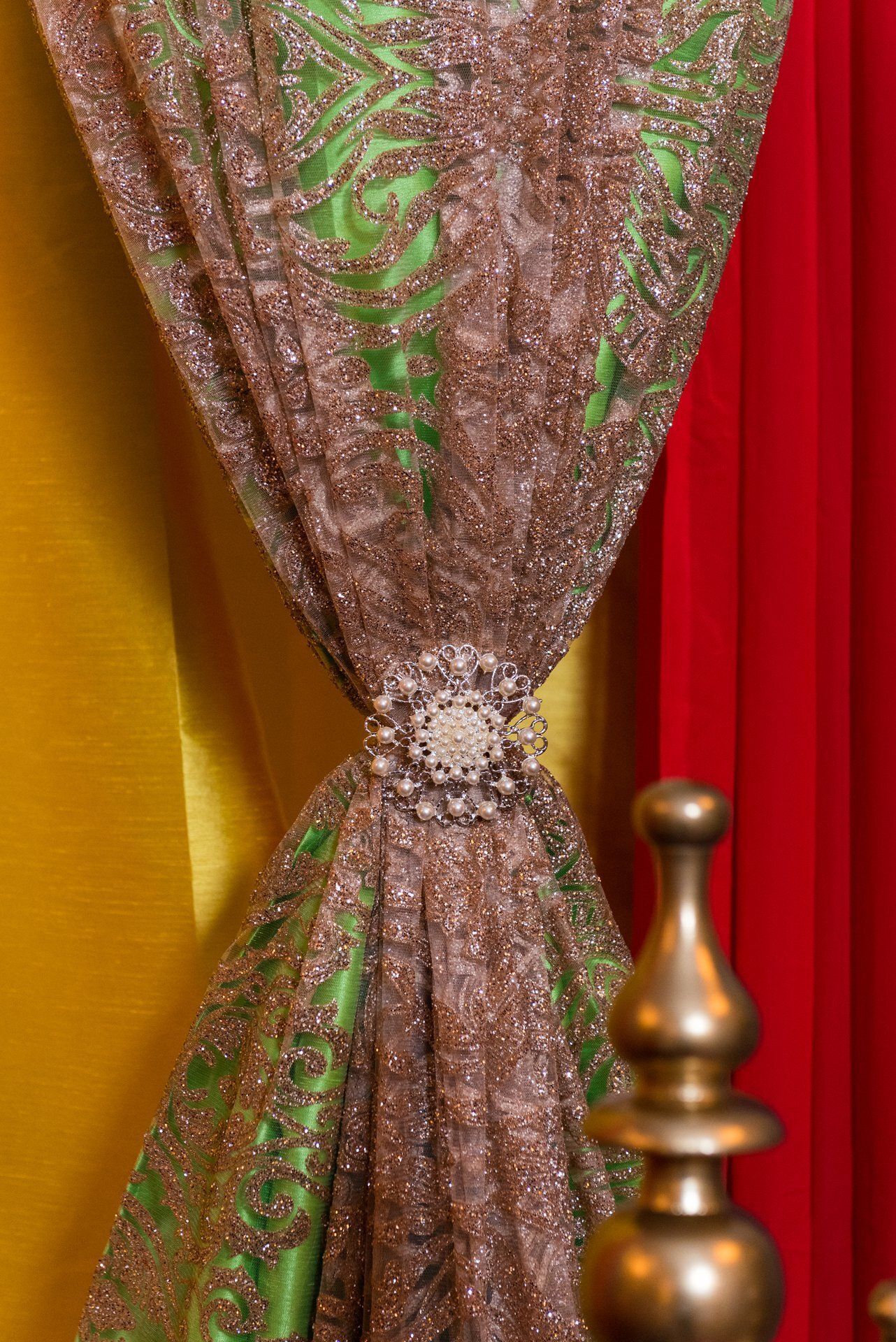 Sheer gold curtain tied back with a sparkling center, beside red and yellow drapes and a brass finial