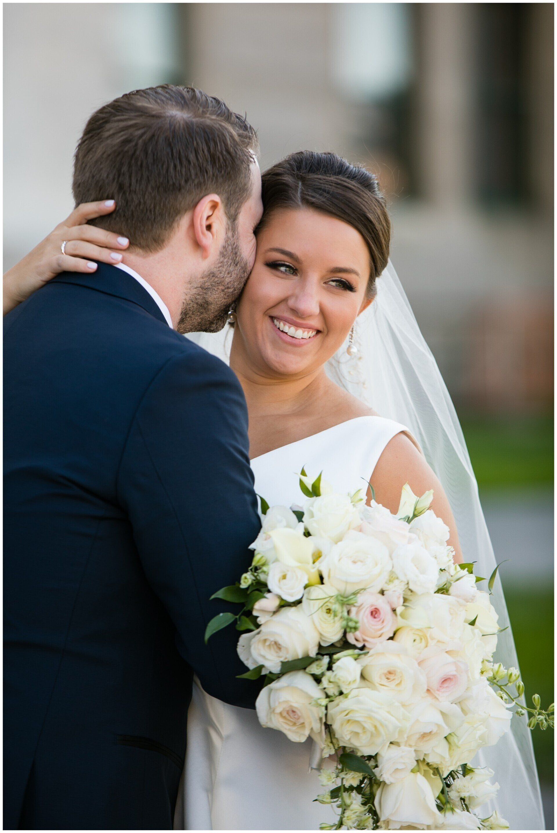 Bride smiling while holding a white bouquet, standing beside the groom outdoors.