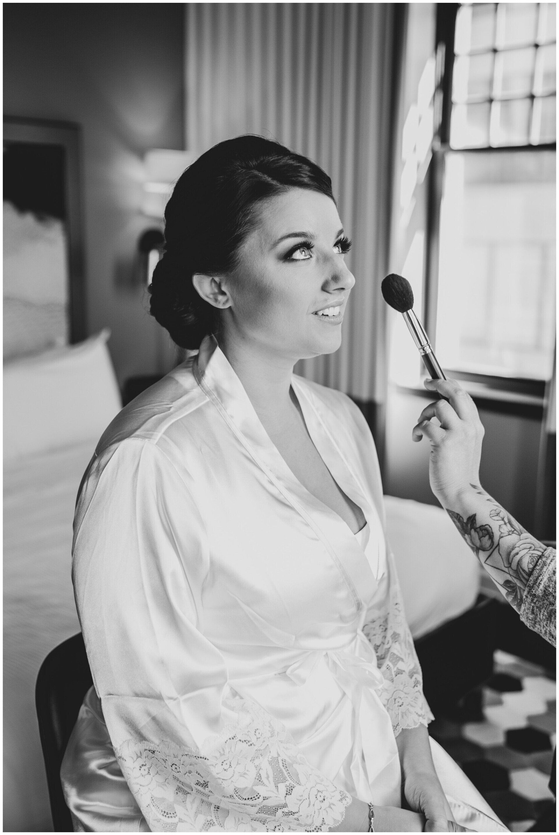Bride having makeup applied by a makeup brush near a window in a black-and-white portrait