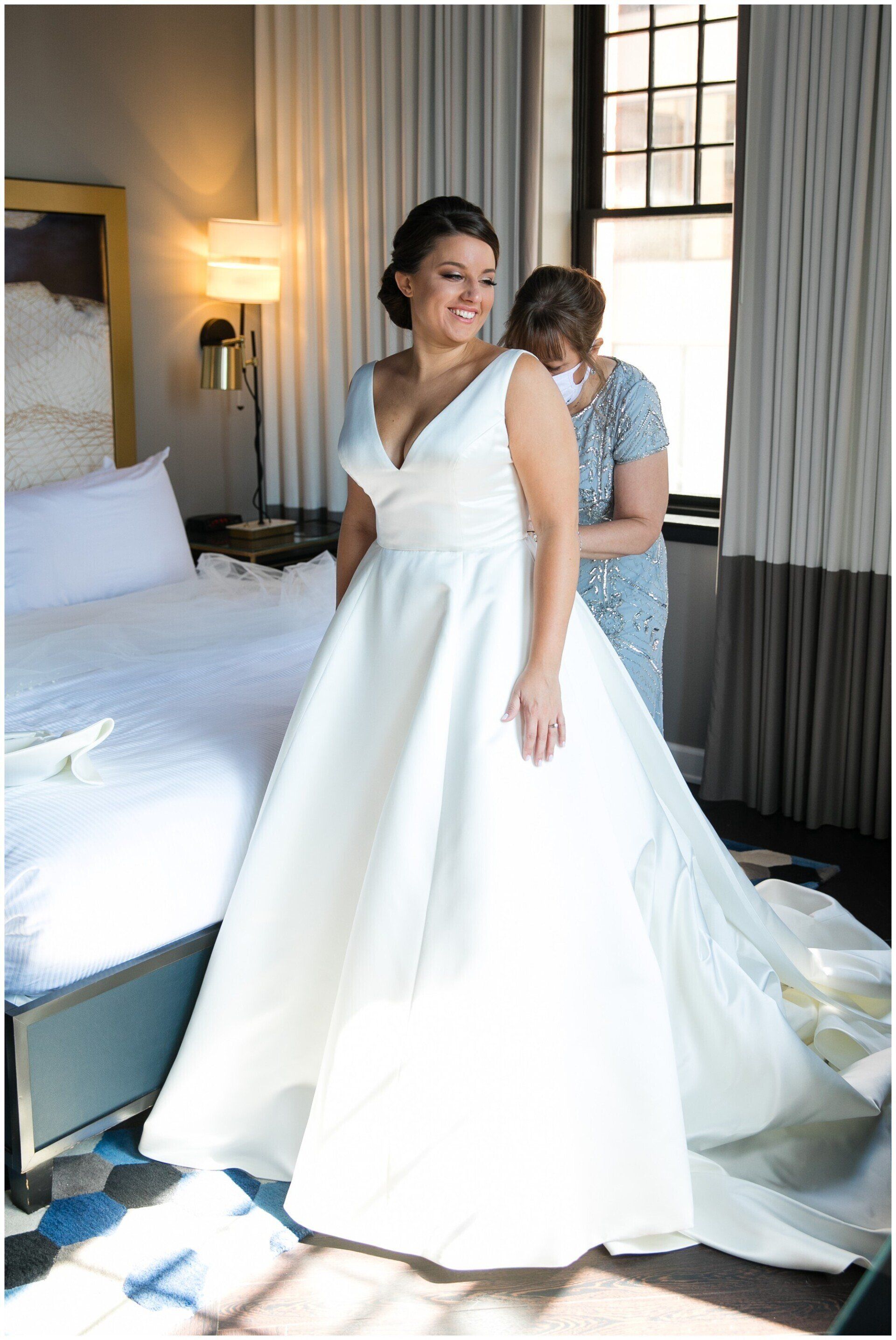 Bride in a white wedding dress smiling in a hotel room, with a train arranged behind her.