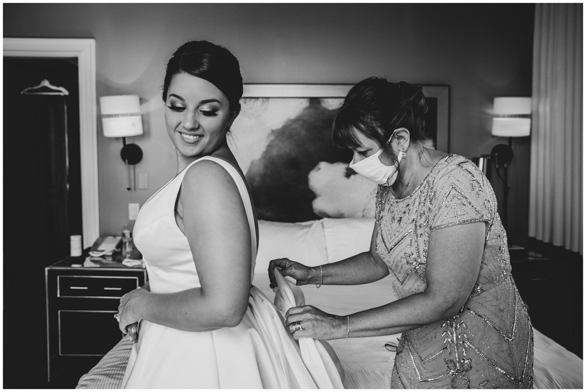 Woman adjusting another woman's wedding dress in a hotel room, both smiling.