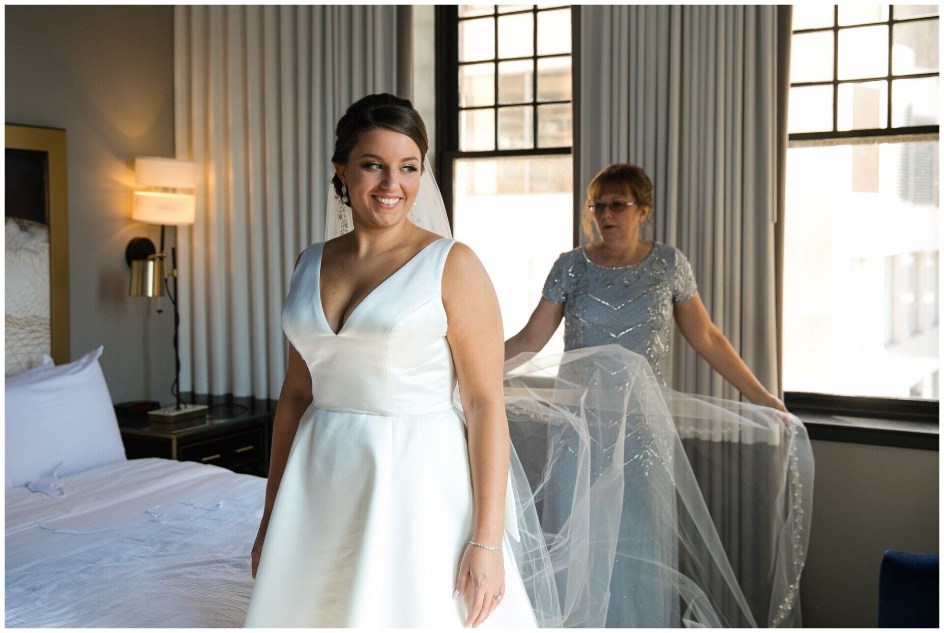 Bride in white dress smiles in a bright room while another woman adjusts her veil behind her.
