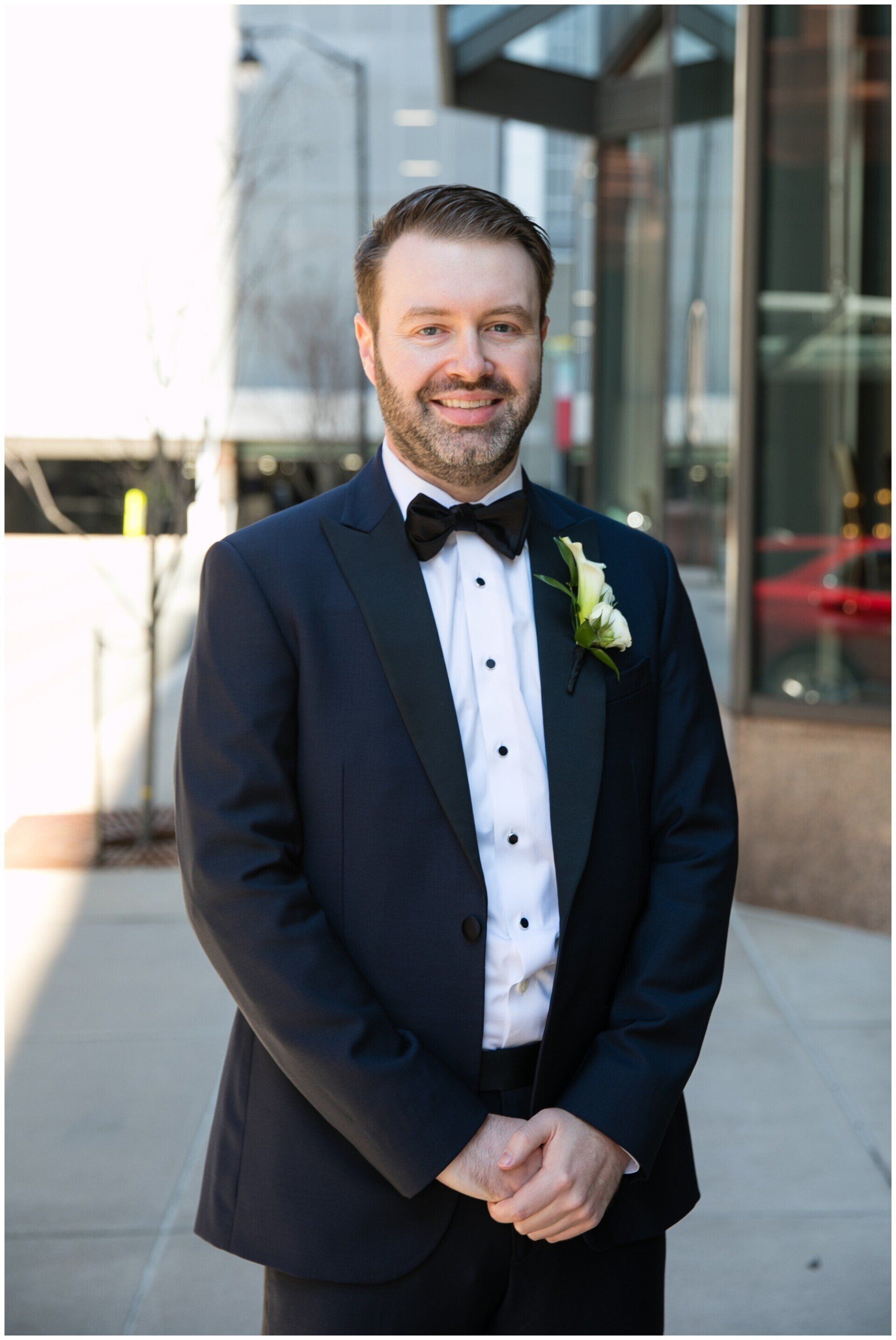 Man in a navy tuxedo with a bow tie standing outdoors beside a building, smiling at the camera