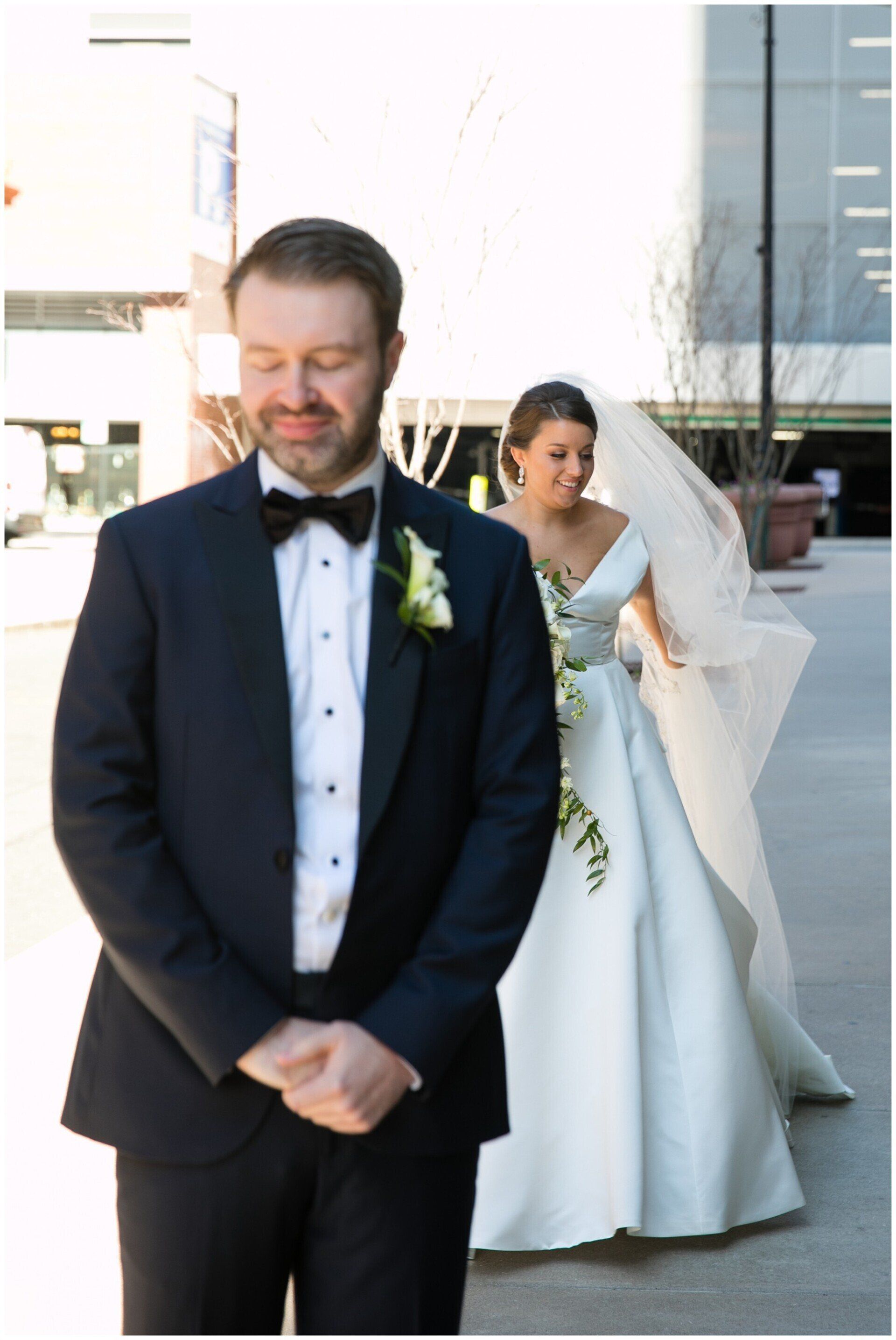 Bride and groom in wedding attire, standing outdoors; groom in dark suit in foreground, bride in white dress behind.