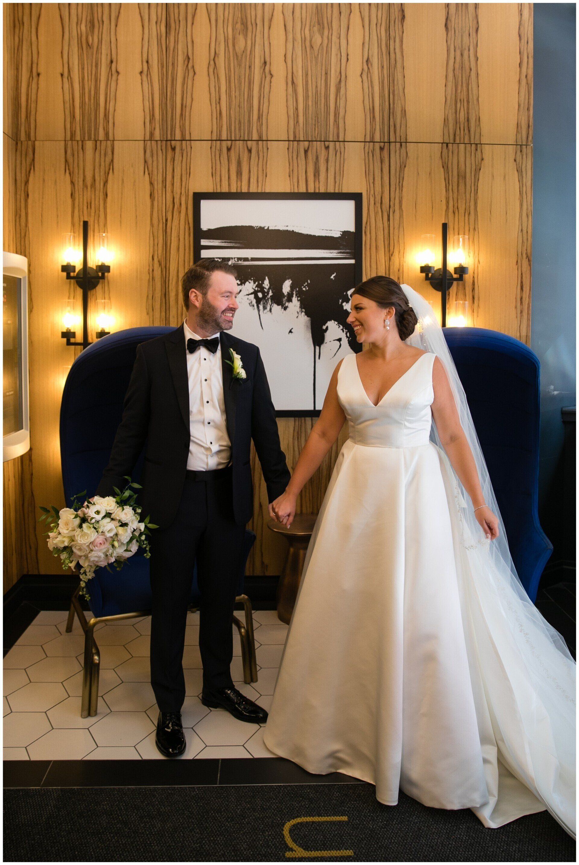 Bride and groom smiling in a wood-paneled room, standing beside blue chairs and wall sconces.