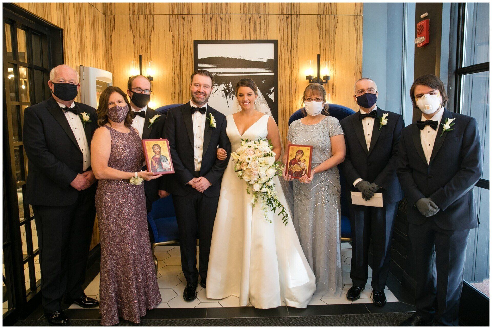 Wedding party posing indoors, bride in white gown holding bouquet, surrounded by guests in formal attire