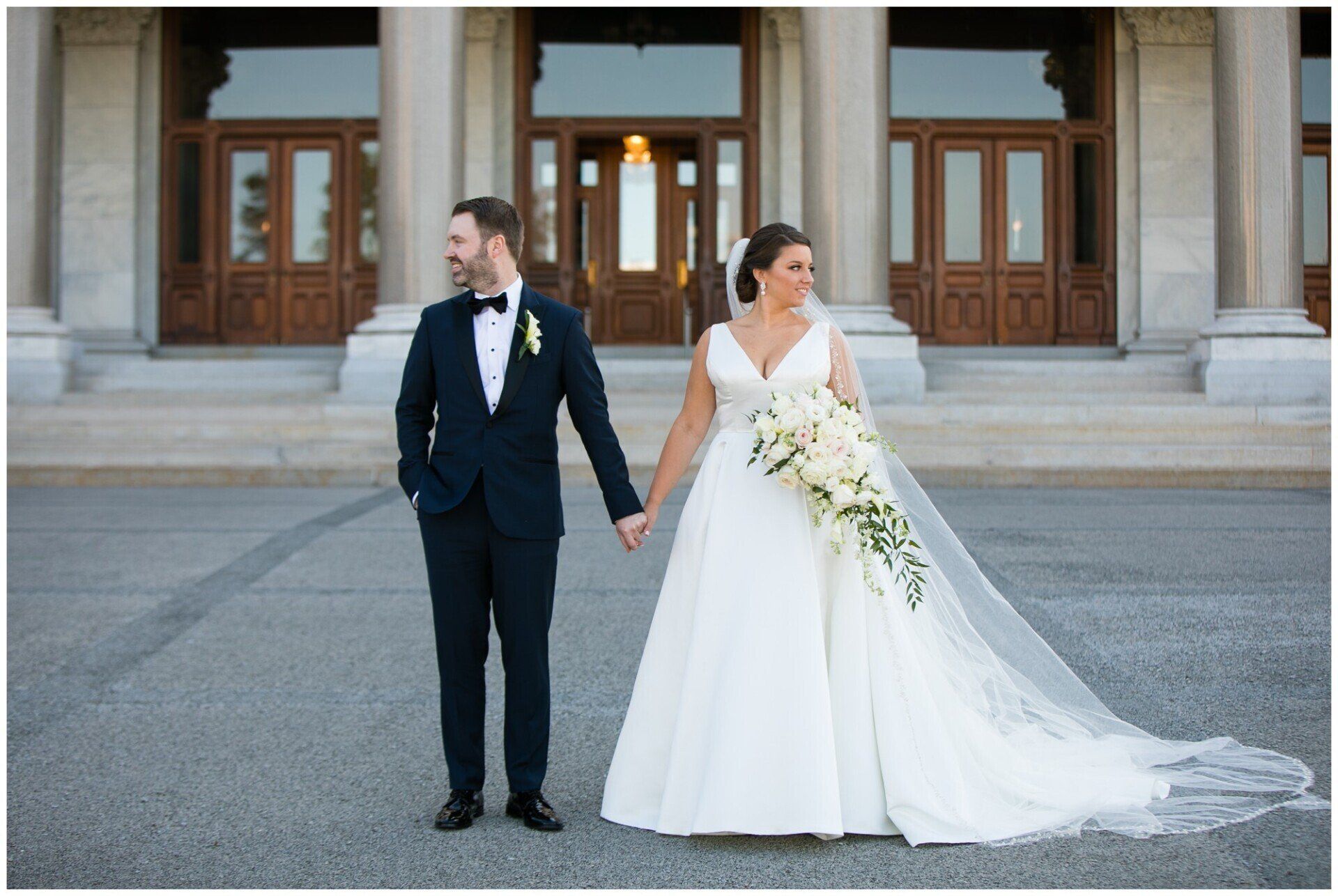 Bride and groom holding hands outside a stone building, the bride in a white gown with a long train.
