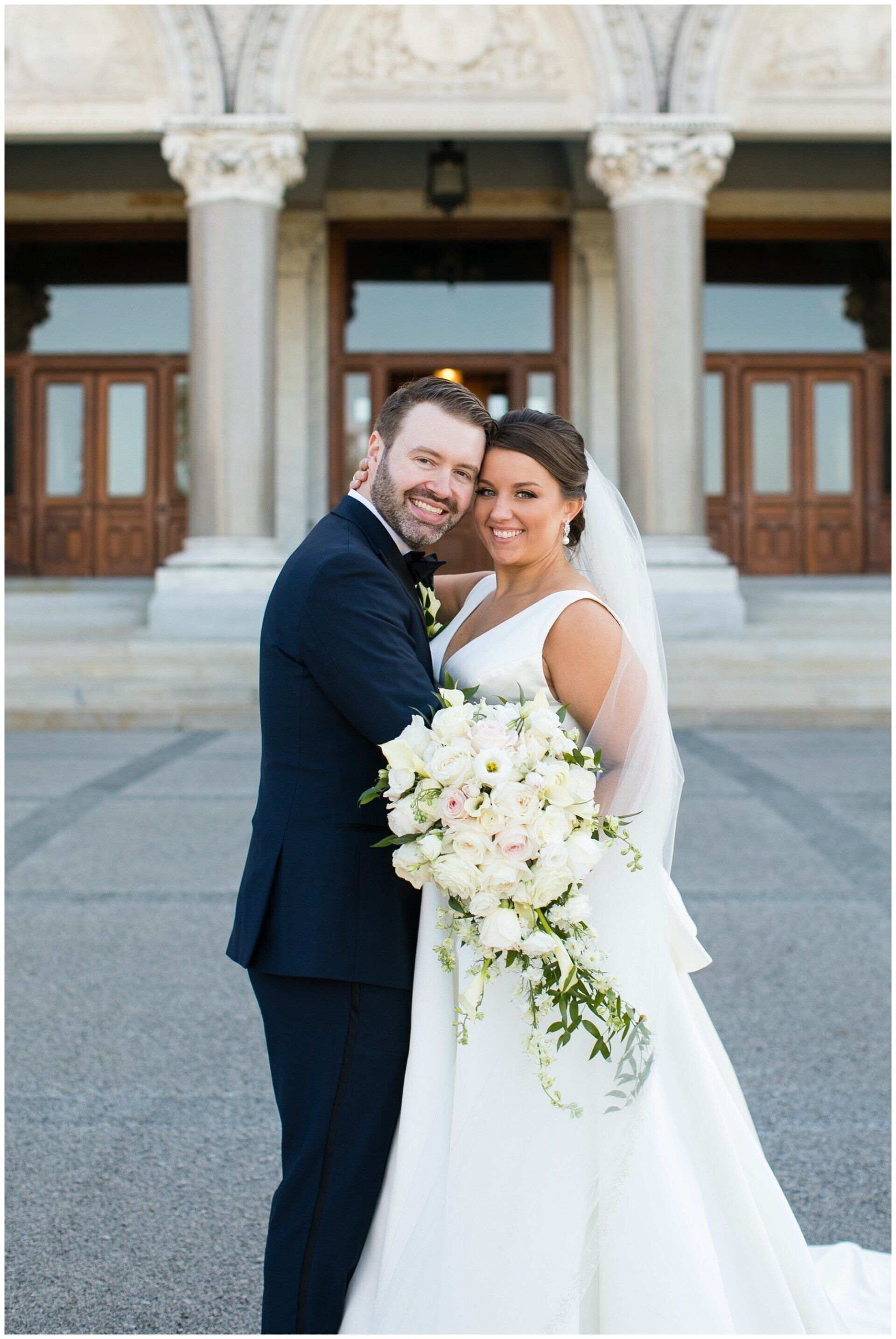 Newlyweds embracing outside a stone building, bride holding a white bouquet