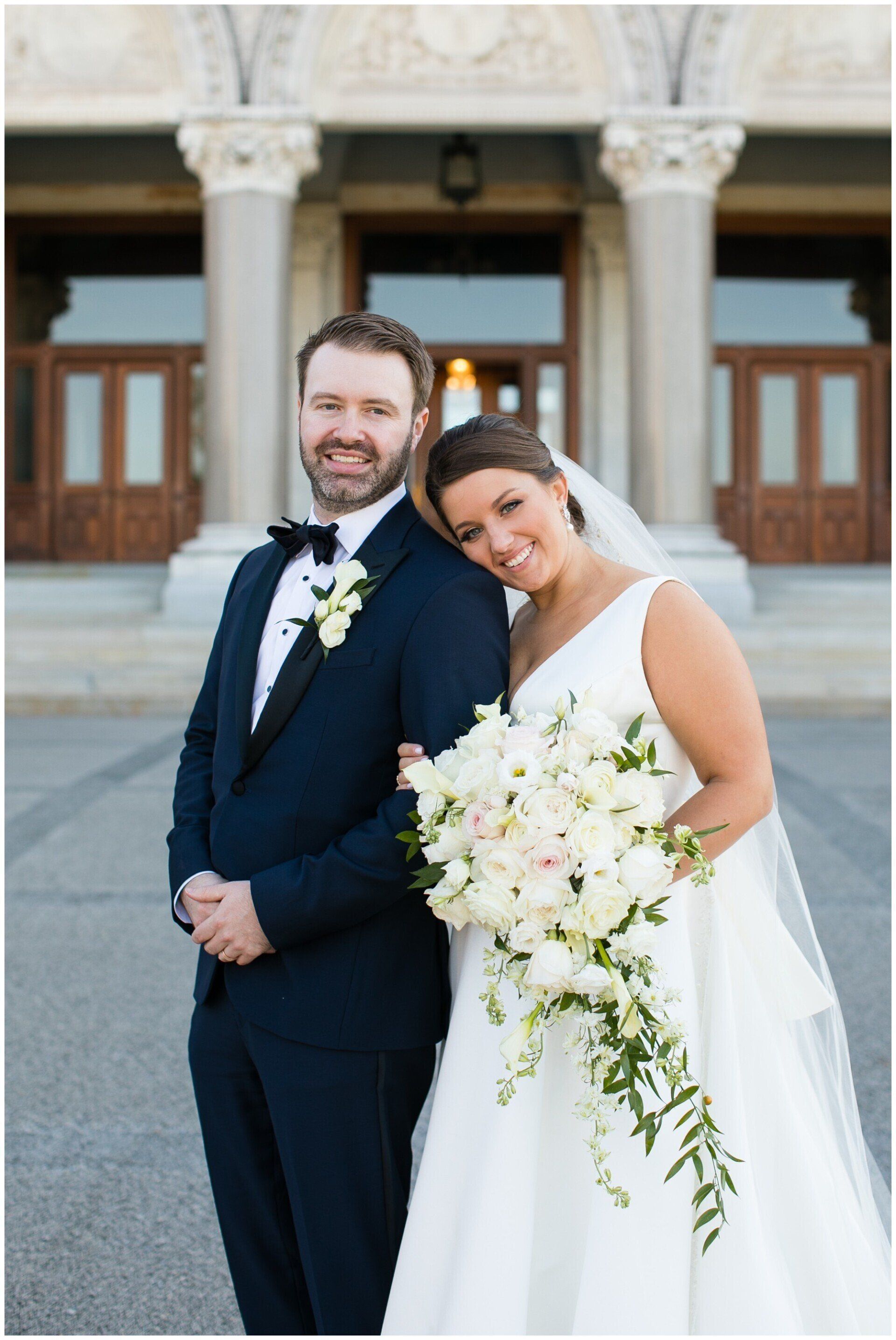 Bride and groom posing outside a building, bride holding a white bouquet and leaning on groom's shoulder.