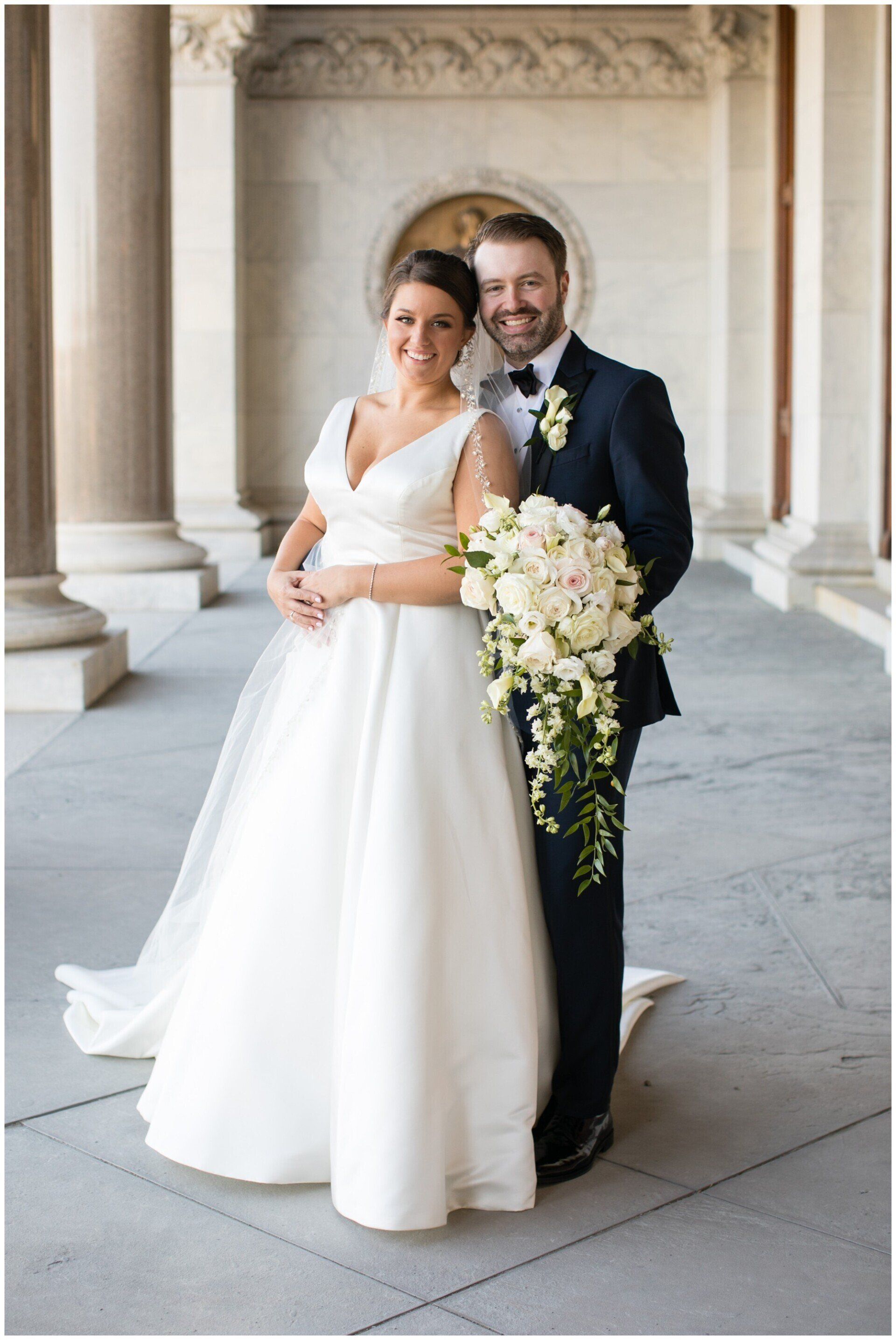 Bride and groom posing in a stone colonnade, holding a bouquet of white flowers