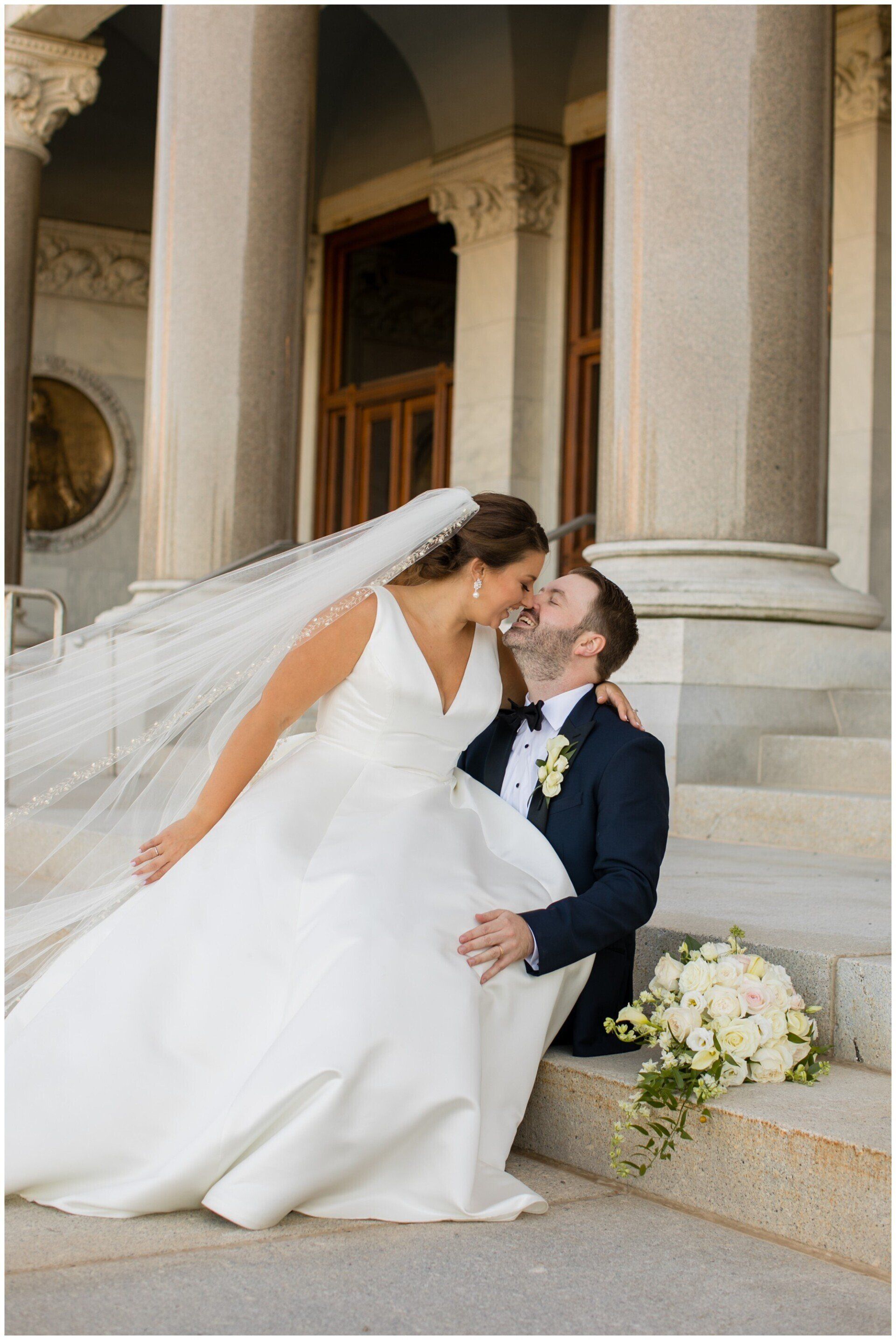Bride and groom kissing on courthouse steps, bride in white gown and veil, bouquet nearby
