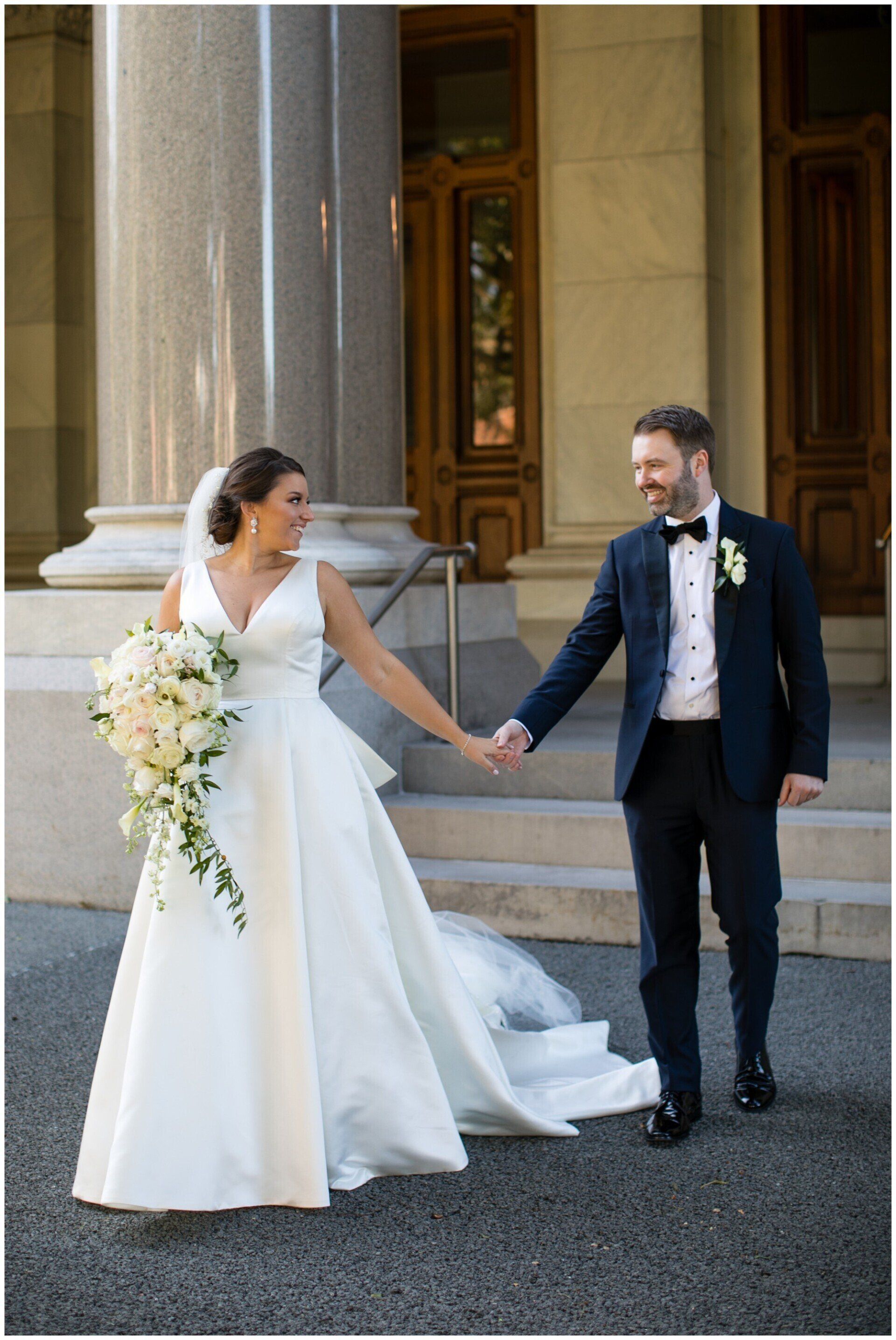Bride and groom holding hands outside a grand building, bride’s veil and bouquet visible