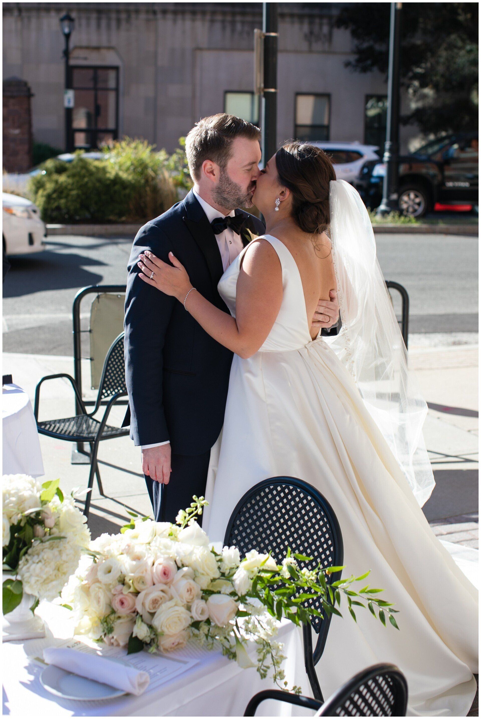 Couple in wedding attire kissing at an outdoor reception table with white flowers and black chairs
