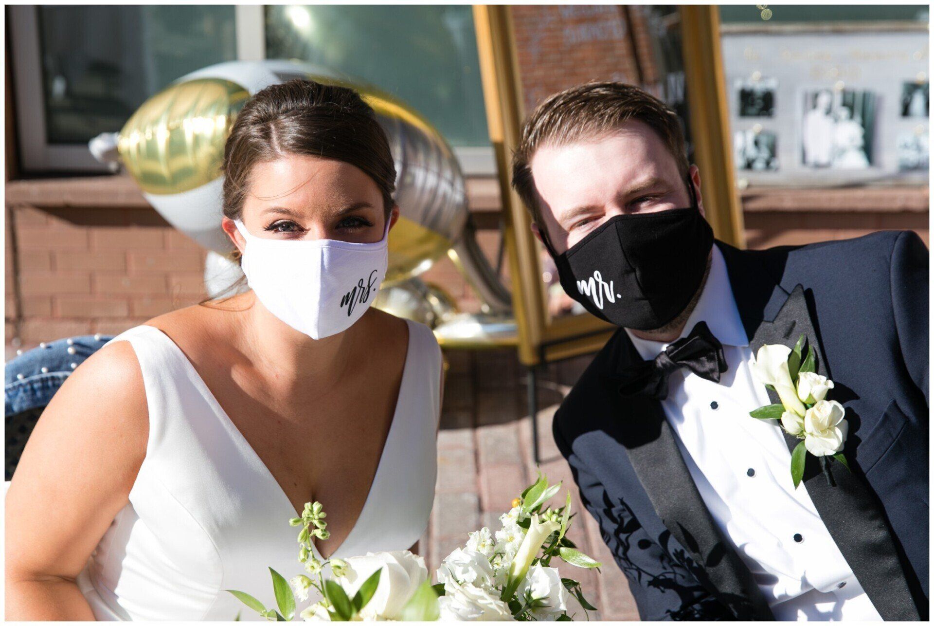 Two people in wedding attire wearing face masks, seated outdoors with flowers.