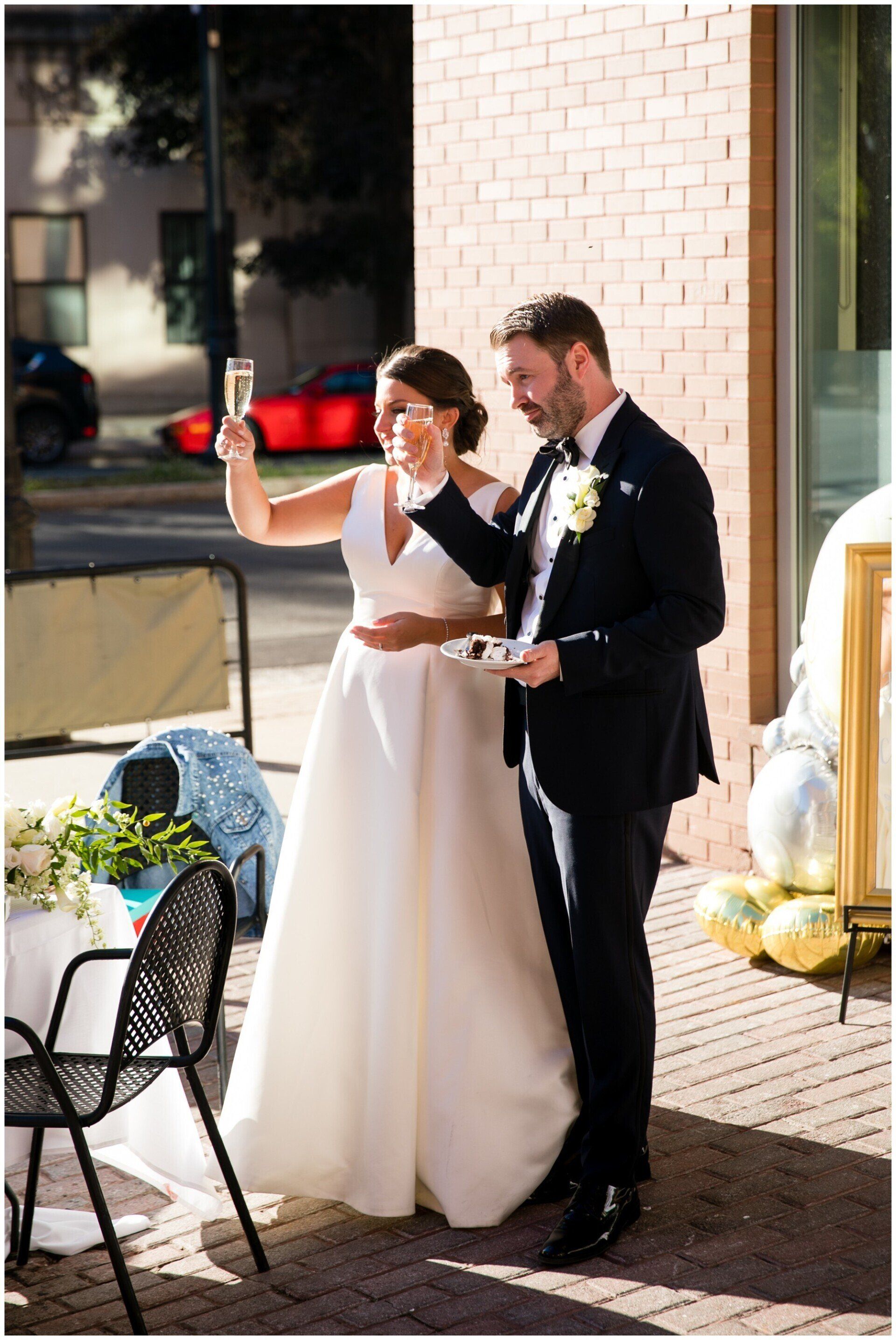 Bride and groom stand outdoors, raising drinks during a wedding toast beside a decorated table.
