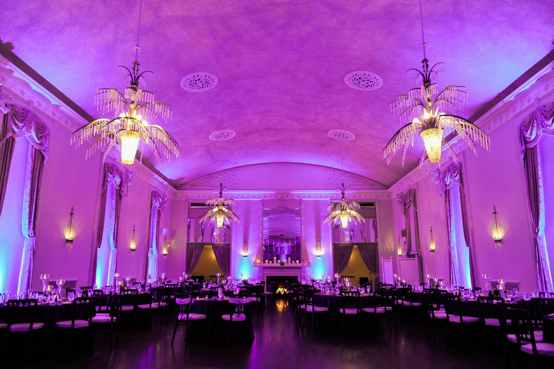 Lavishly lit banquet hall with chandeliers and purple ambient lighting, set with round tables.