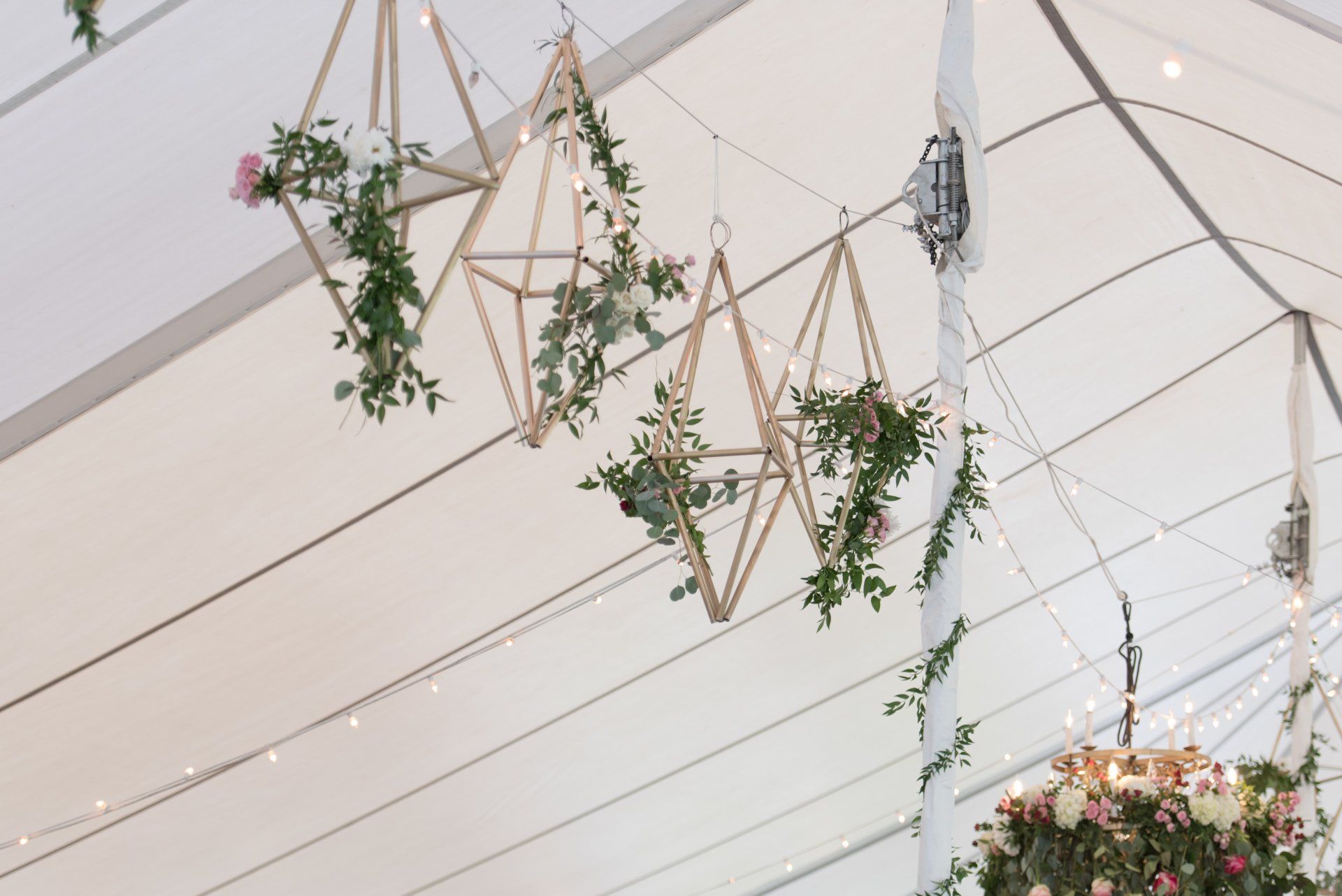 Hanging wooden geometric wreaths with greenery on a white tent ceiling, lit by string lights