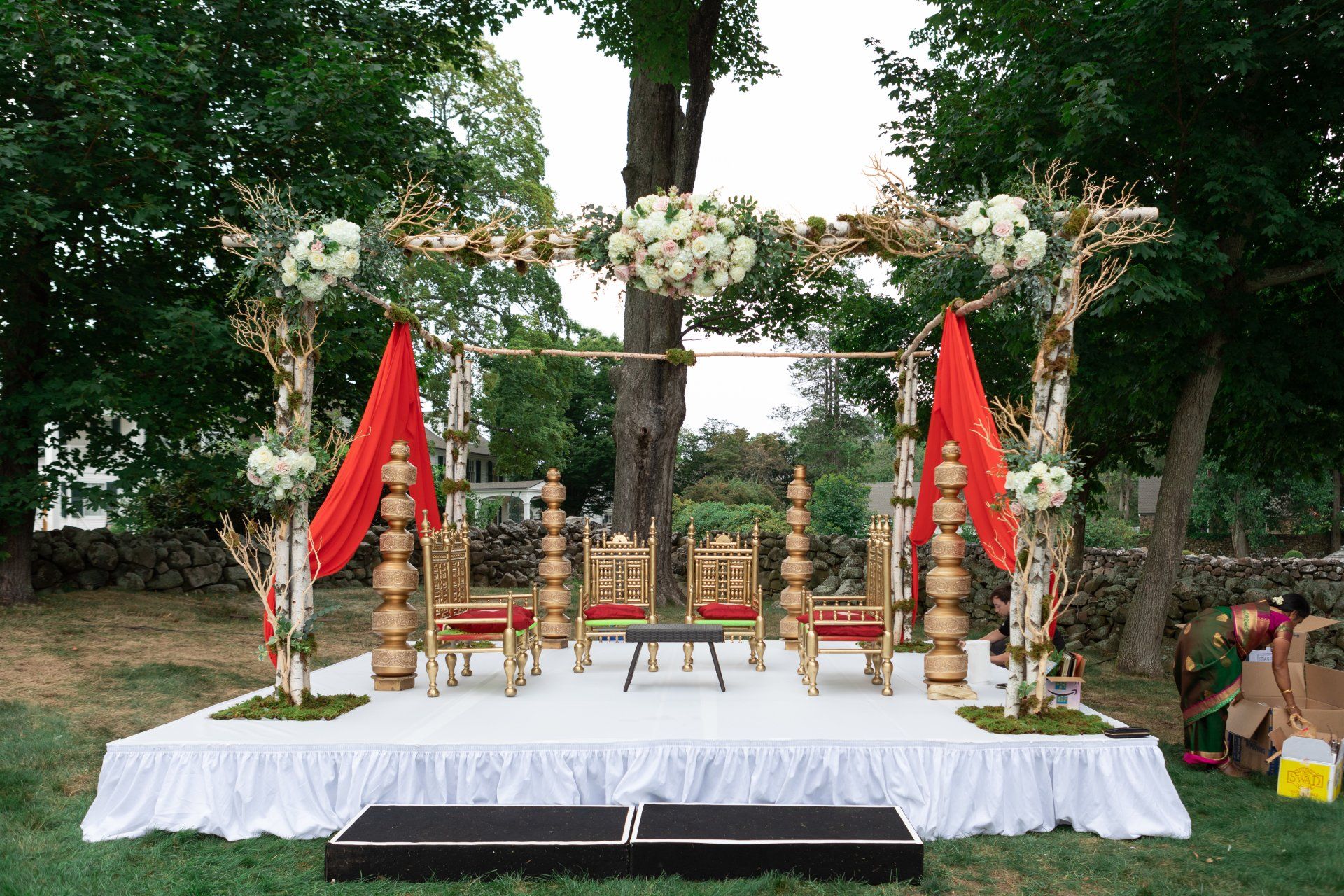 Outdoor wedding altar with white flowers, red flags, and gold chairs on a white stage under trees