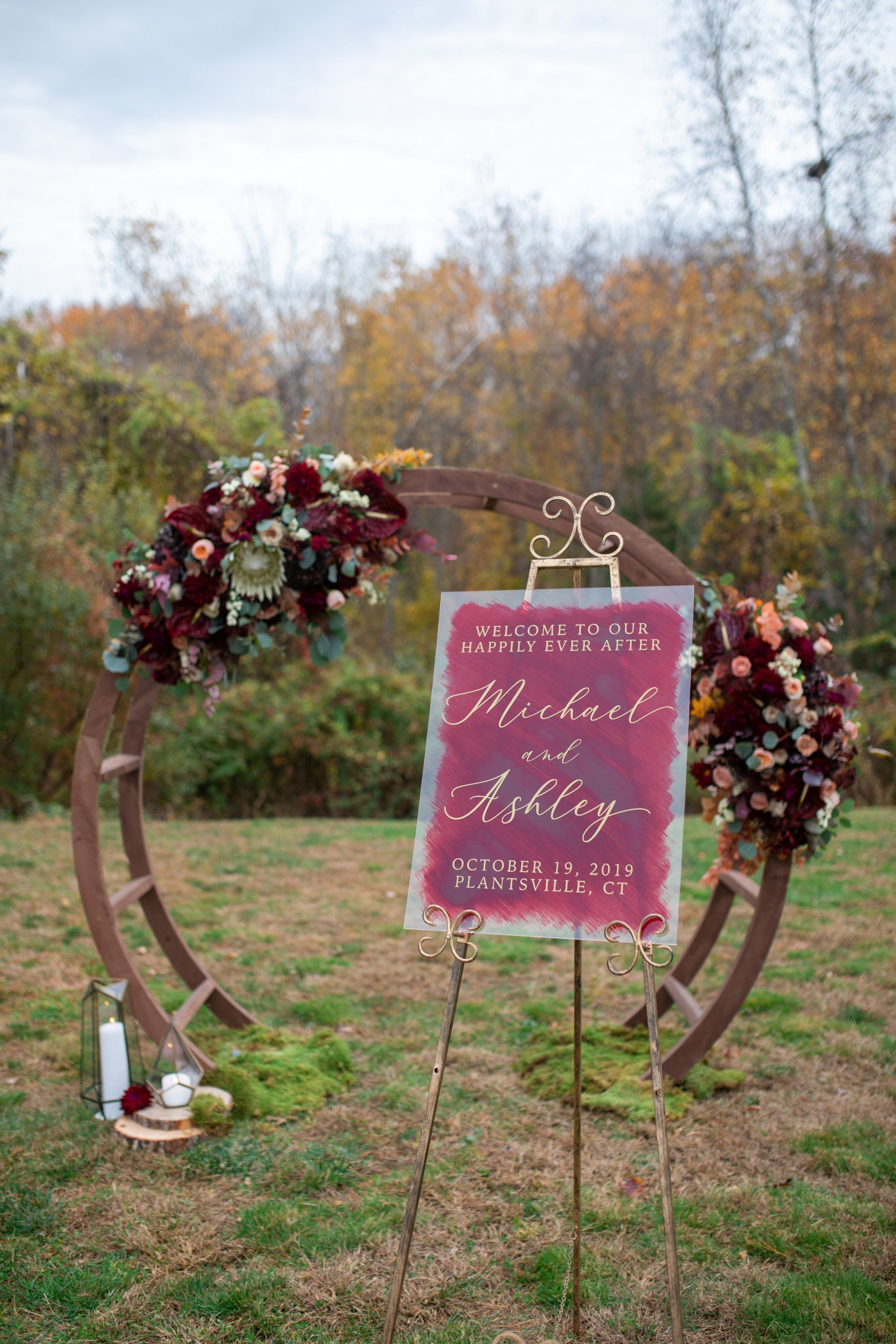Rustic outdoor wedding welcome sign with burgundy flowers and wooden hoops on grass.