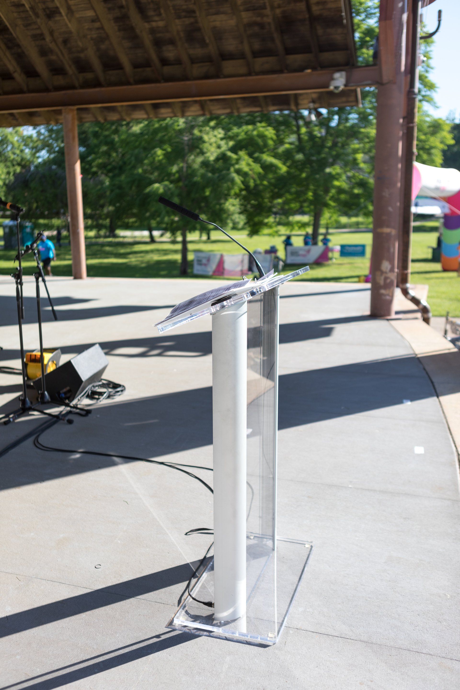 Two silver microphone stands on an outdoor stage under a pavilion, with trees and grass in the background.