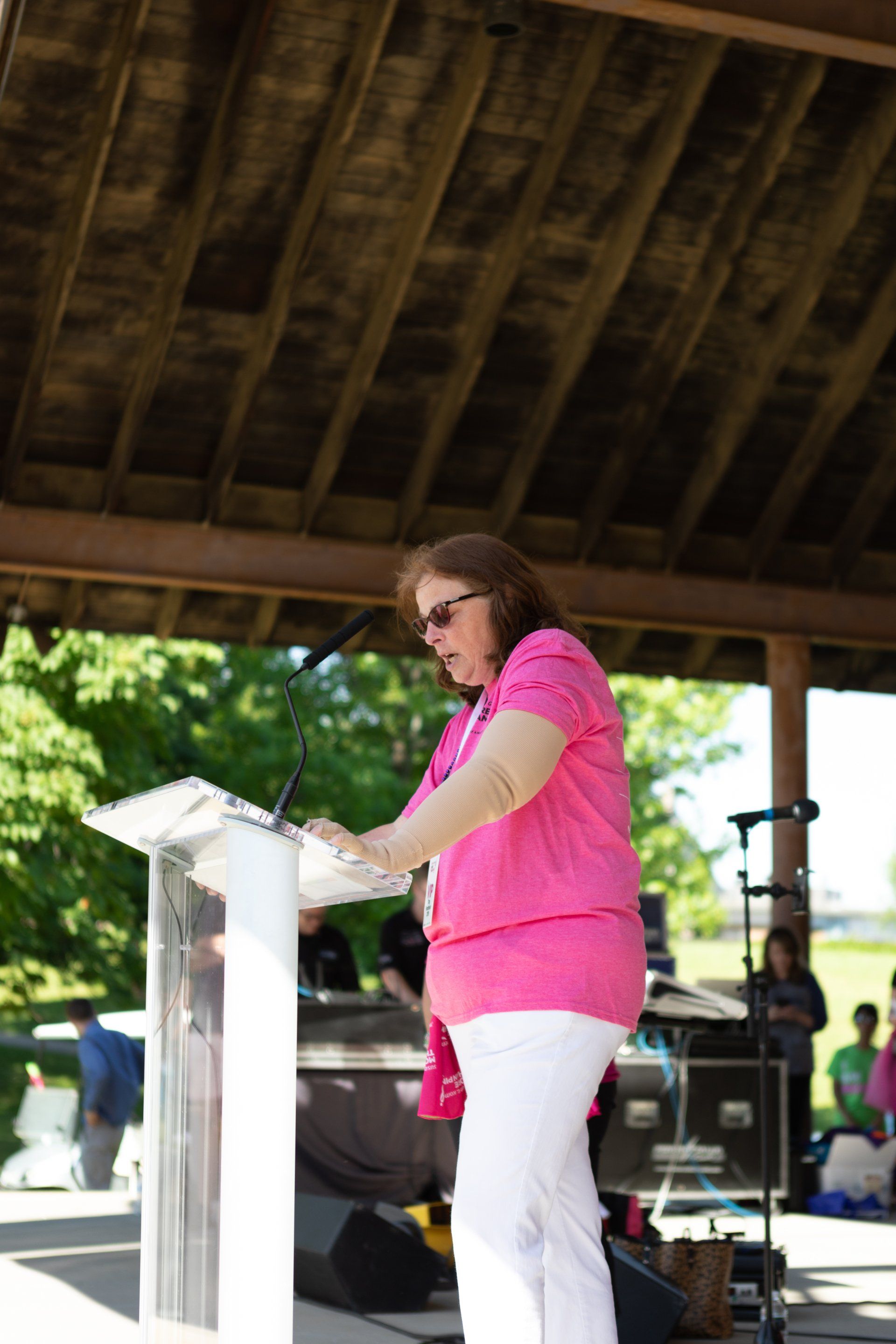 Woman in pink shirt speaking at a podium under a pavilion at an outdoor event